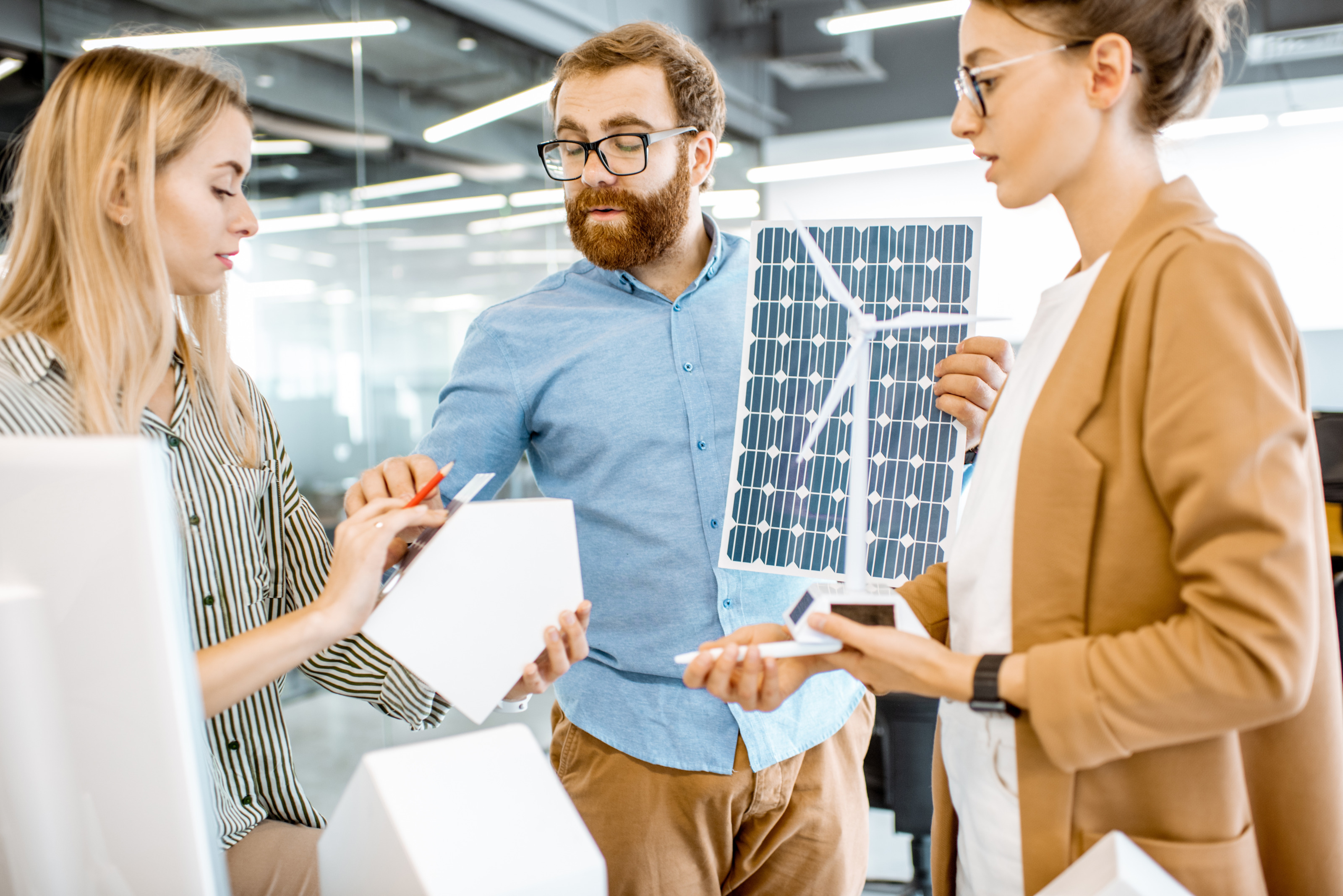 Drei Personen in Gespr&auml;ch, eine Person h&auml;lt Modell eines Windrades, ein Person h&auml;lt Photovoltaikfolie und eine weitere h&auml;lt Modell eines Hauses, auf dem sie Folie appliziert