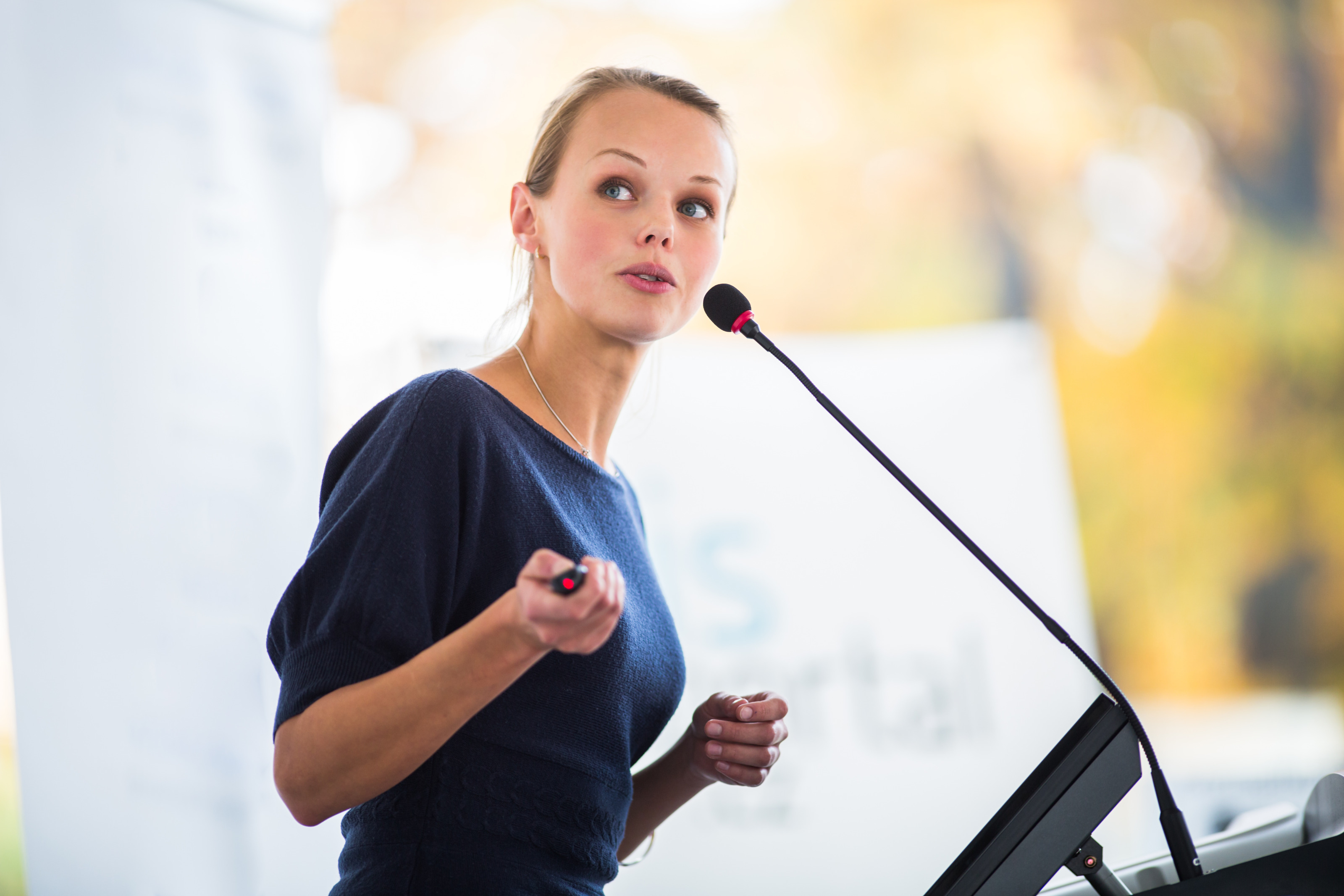 Seitliche Aufnahme einer Person, die an einem Pult steht und in ein Mikrofon spricht. Die Person h&auml;lt eine Fernbedienung in der rechten Hand. Der Hintergrund ist verschwommen