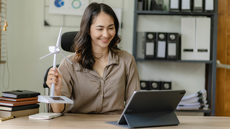 Person an Schreibtisch sitzend mit Modell eines Windrades in Hand blickt auf vor ihr stehendes Tablet
