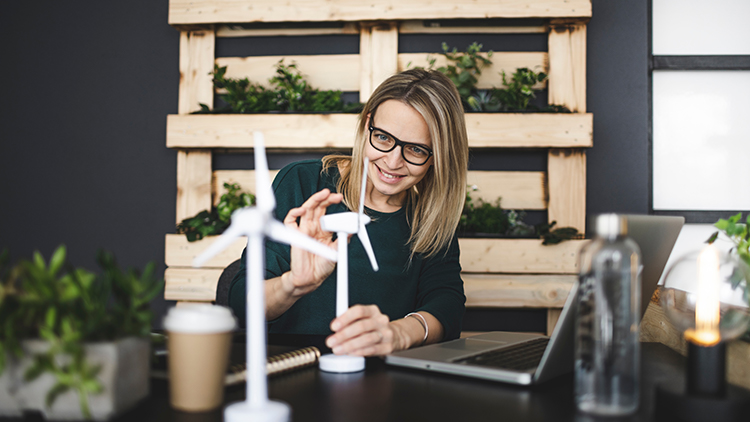 Person mit Brillen sitzt lächelnd an Schreibtisch und blickt auf Modelle von Windrädern, vor ihr aufgeklappter Laptop