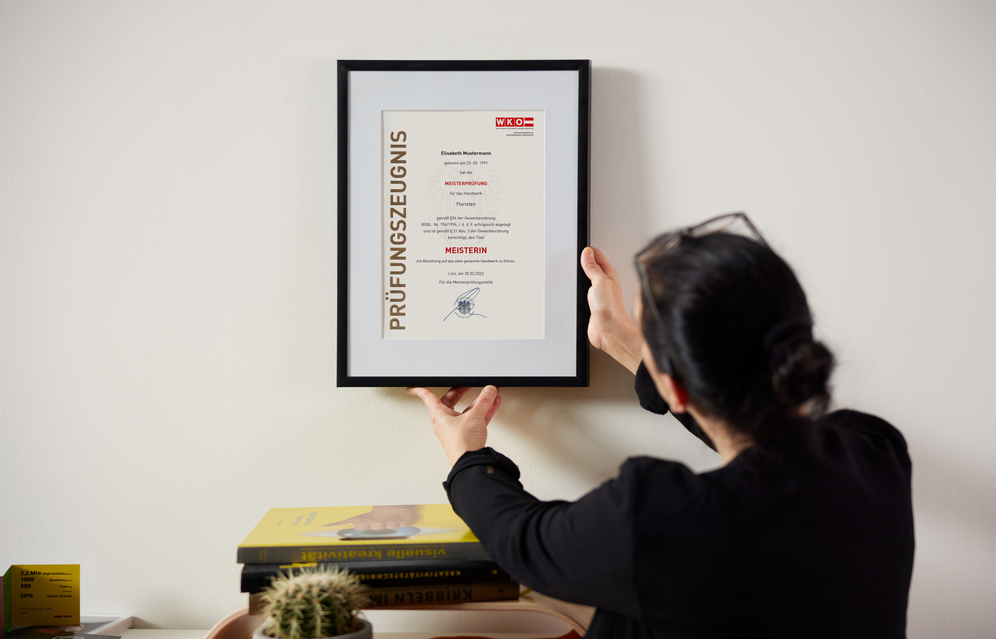 Person mit dunklen geschlossenen Haaren, dunkler Kleidung und Brille steht vor einer Wand und h&auml;lt einen Rahmen mit Passepartout und Pr&uuml;fungszeugnis Meisterin in H&auml;nden, daneben steht eine Pflanze sowie liegen B&uuml;cher auf einem Schrank