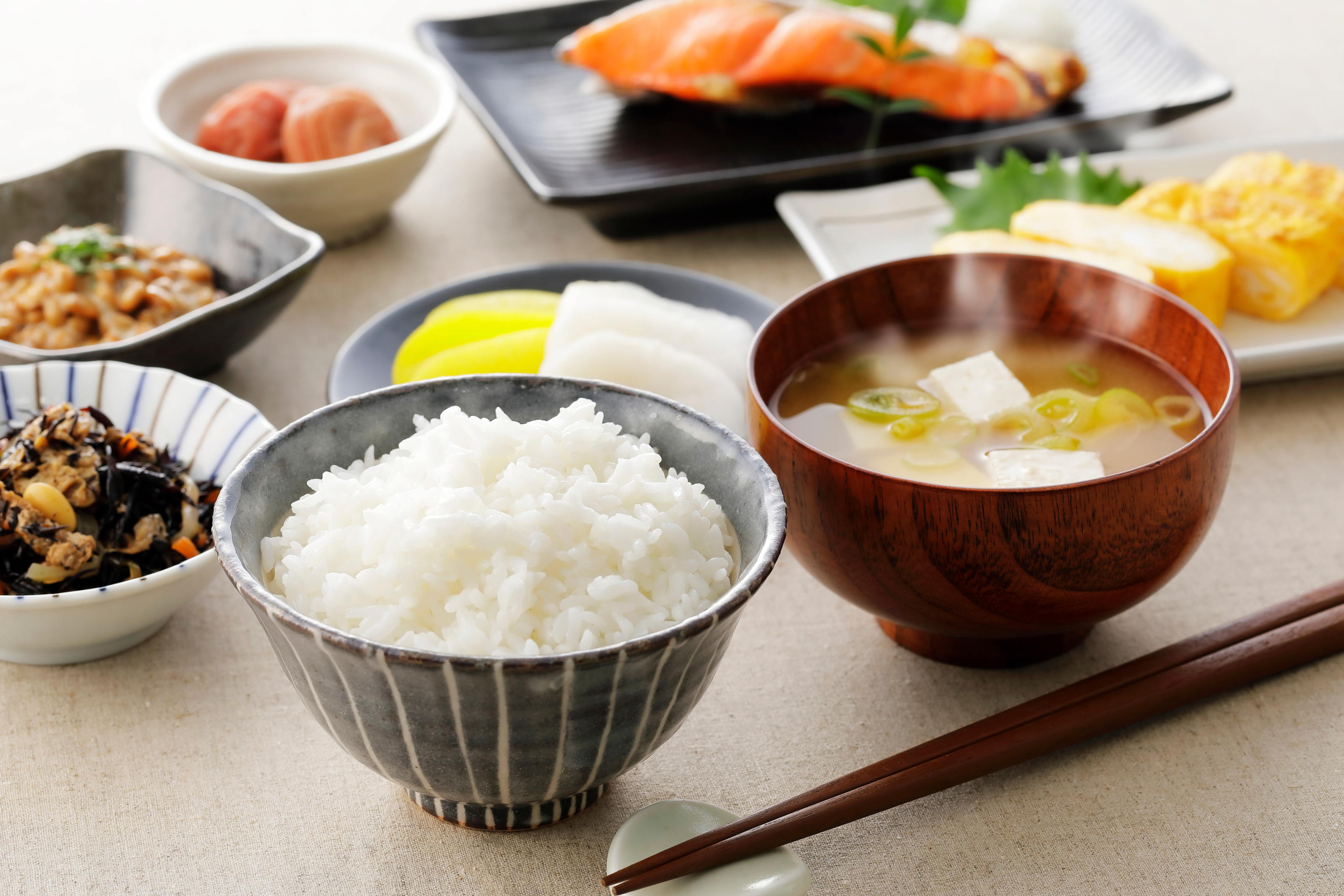 Verschiedene Schälchen und Teller gefüllt mit japanischen Frühstücksspeisen auf einem beigen Untergrund, daneben Stäbchen
