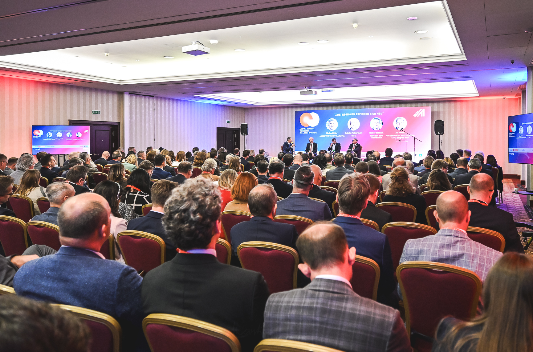 Ein gro&szlig;er Konferenzsaal ist mit zahlreichen Personen besetzt, die auf eine B&uuml;hne am vorderen Ende des Raumes ausgerichtet sind. Auf der B&uuml;hne sitzen mehrere Personen auf einem Podium. Dahinter zeigt eine gro&szlig;formatige Pr&auml;sentationsfl&auml;che Programminhalte mit Fotos und Namen der Vortragenden