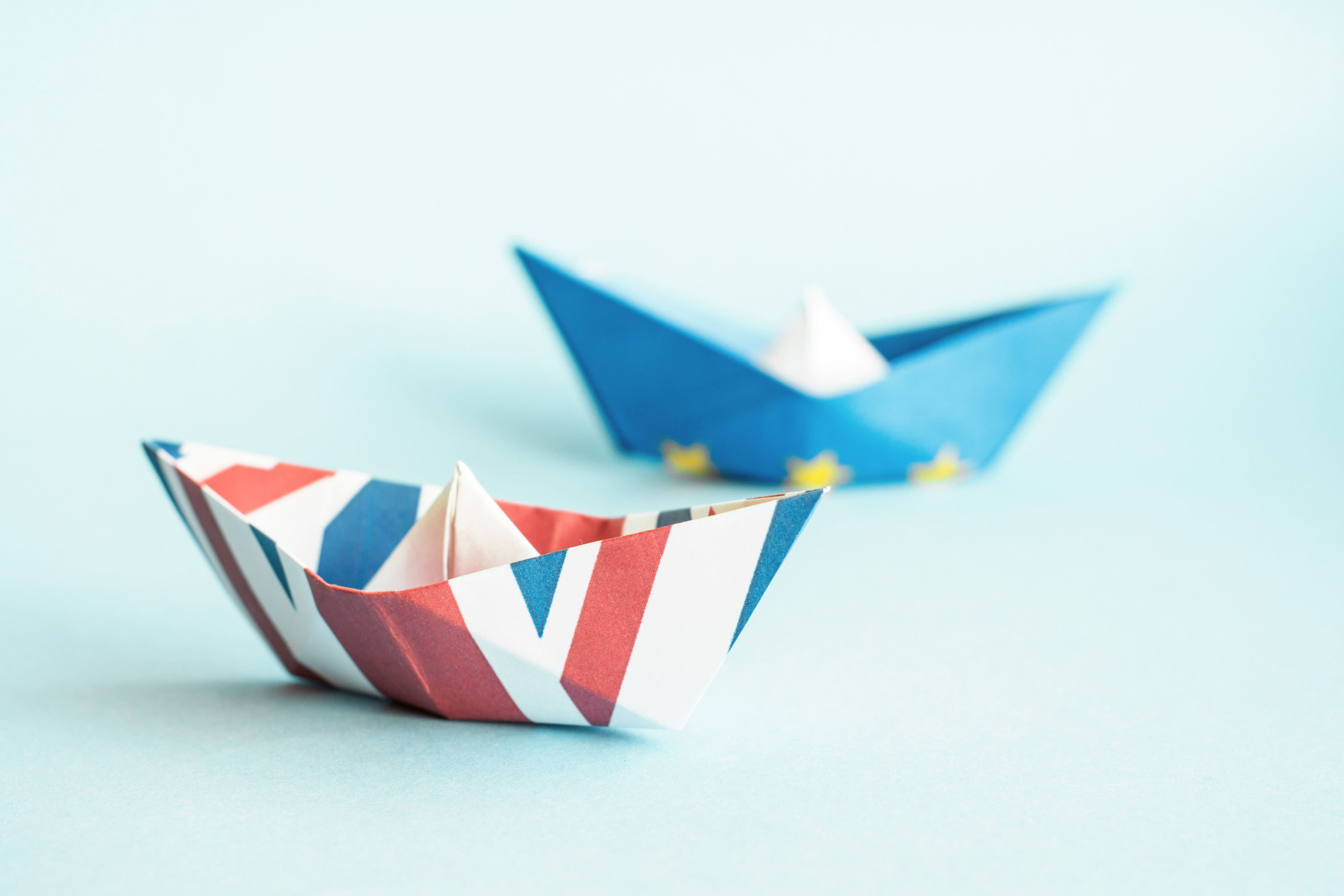 Zwei Papierschiffchen sind auf einem hellblauen Untergrund platziert, das vordere mit United Kingdom Flagge gefaltet und das hintere mit der Symbolik der Europ&auml;ischen Union