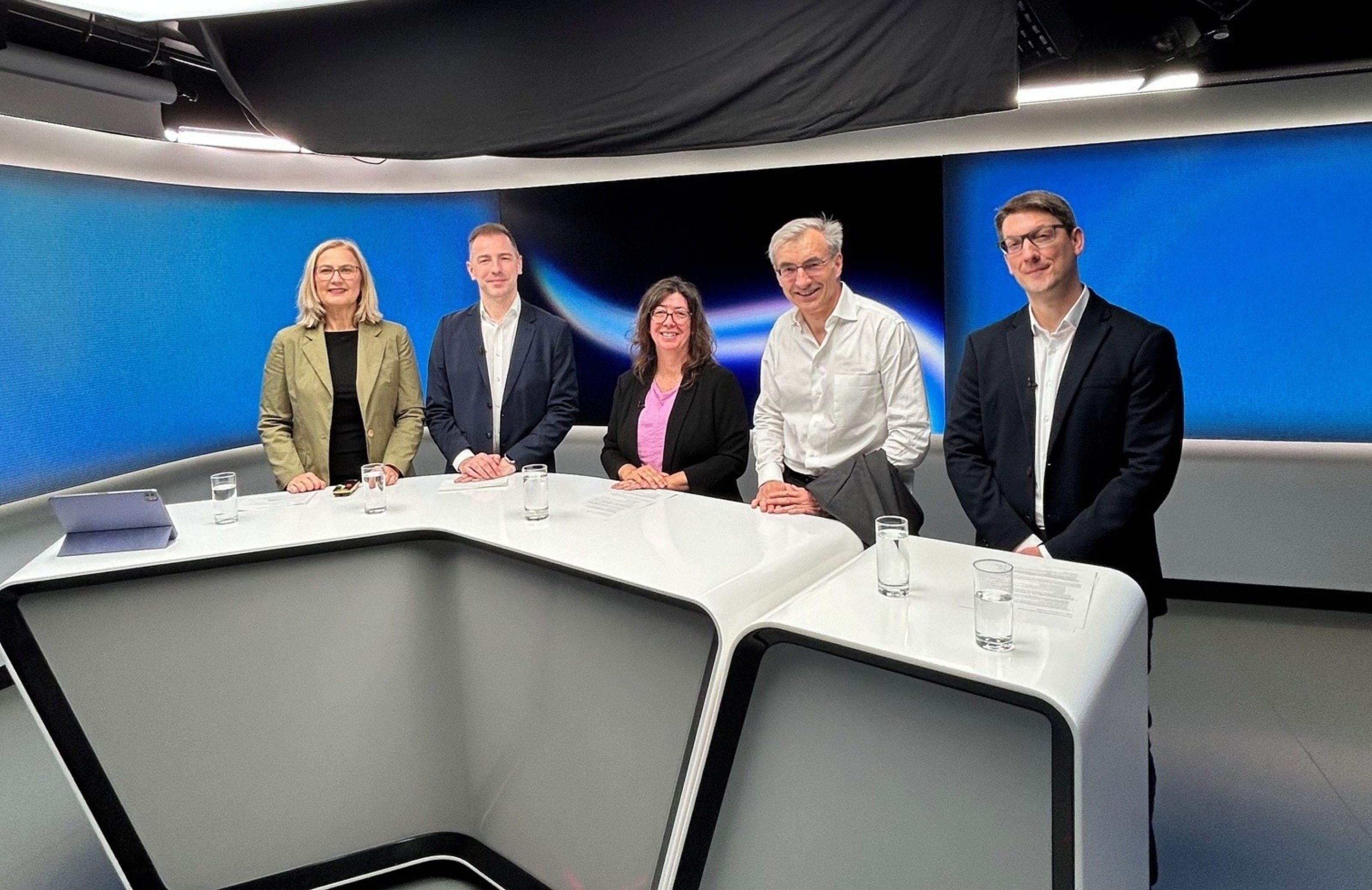 F&uuml;nf Personen stehen in einem modernen TV-Studio hinter einem Studiotisch.