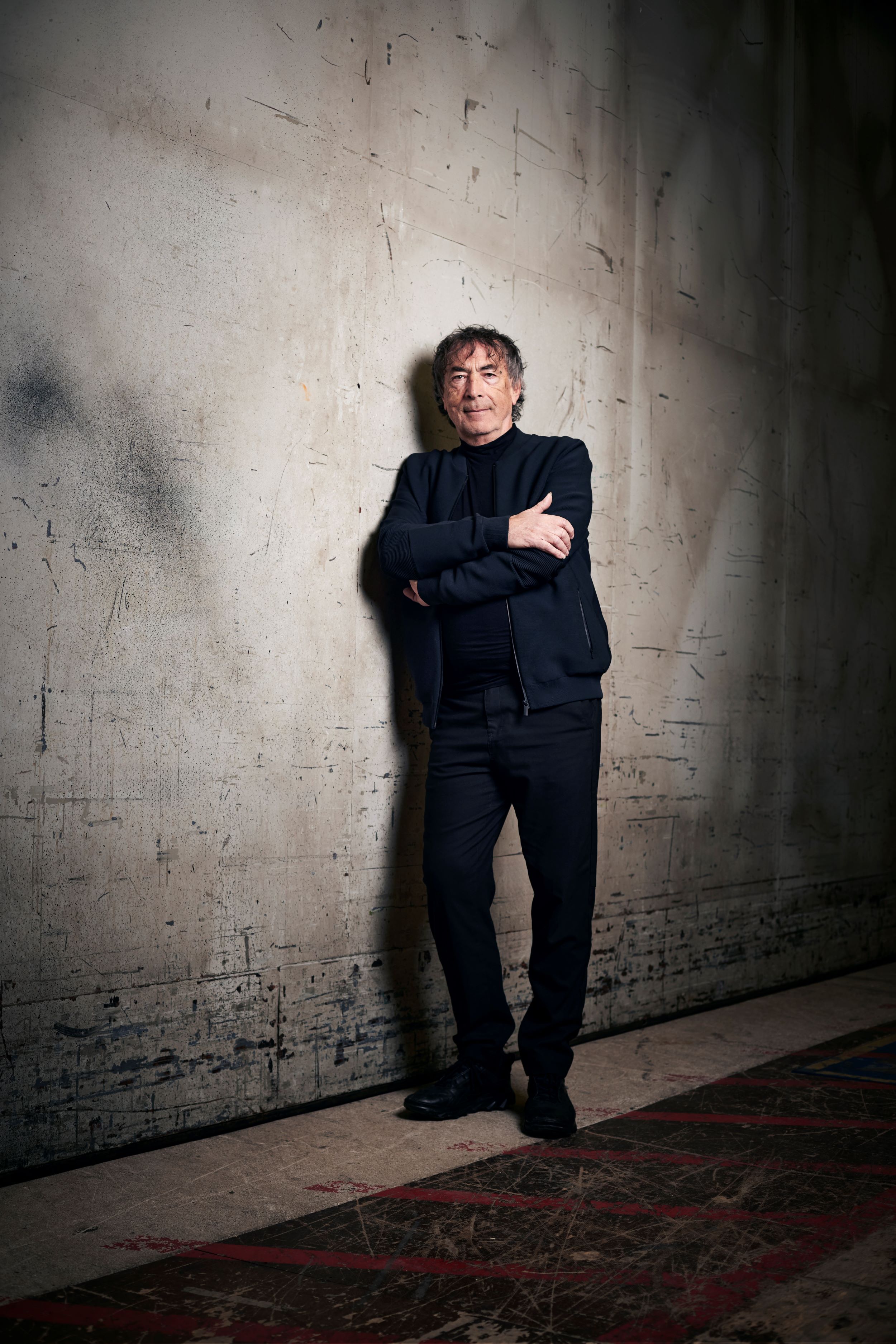 Portr&auml;tfoto: Person in dunkler Kleidung steht mit verschr&auml;nkten Armen vor strukturierter Wand