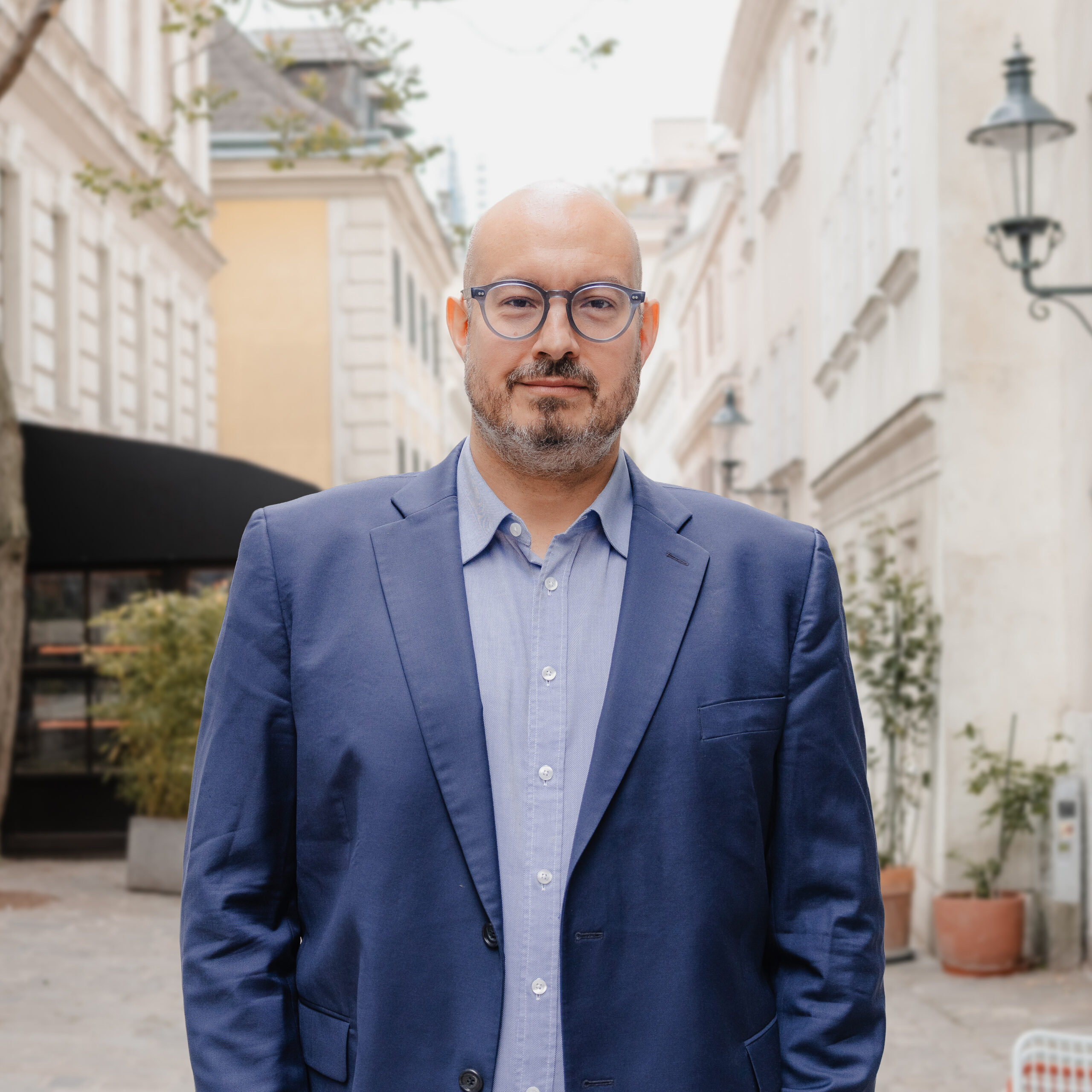 Portr&auml;tfoto: Person mit Brille und blauem Sakko in einer Gasse mit Geb&auml;uden