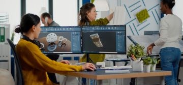 Person mit Kopfh&ouml;rern um den Hals sitz an Schreibtisch vor zwei gro&szlig;en Monitoren auf denen Renderings von Zahnr&auml;dern und Bauteilen zu sehen sind, im Hintergrund weitere Personen stehend in B&uuml;roraum