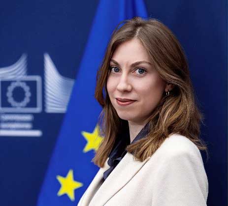 Person im hellen Blazer vor einer EU-Flagge und institutionellem Hintergrund.