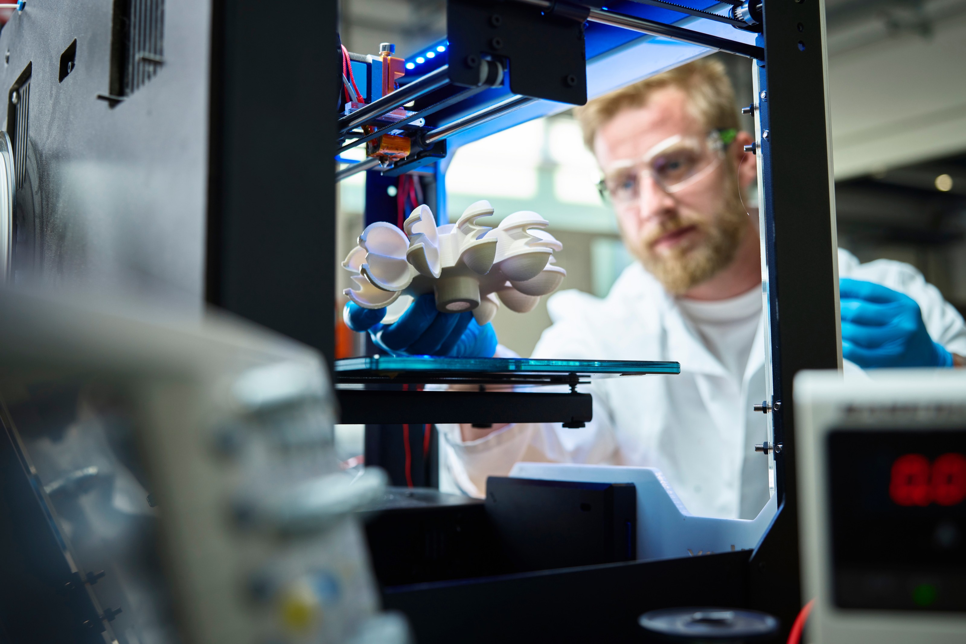 Im Vordergrund steht ein 3D-Drucker, in dessen Mitte eine Platte ist. Neben dem Drucker steht eine Person mit Schutzbrille, Kittel und Handschuhen, die &uuml;ber die Platte ein Objekt h&auml;lt