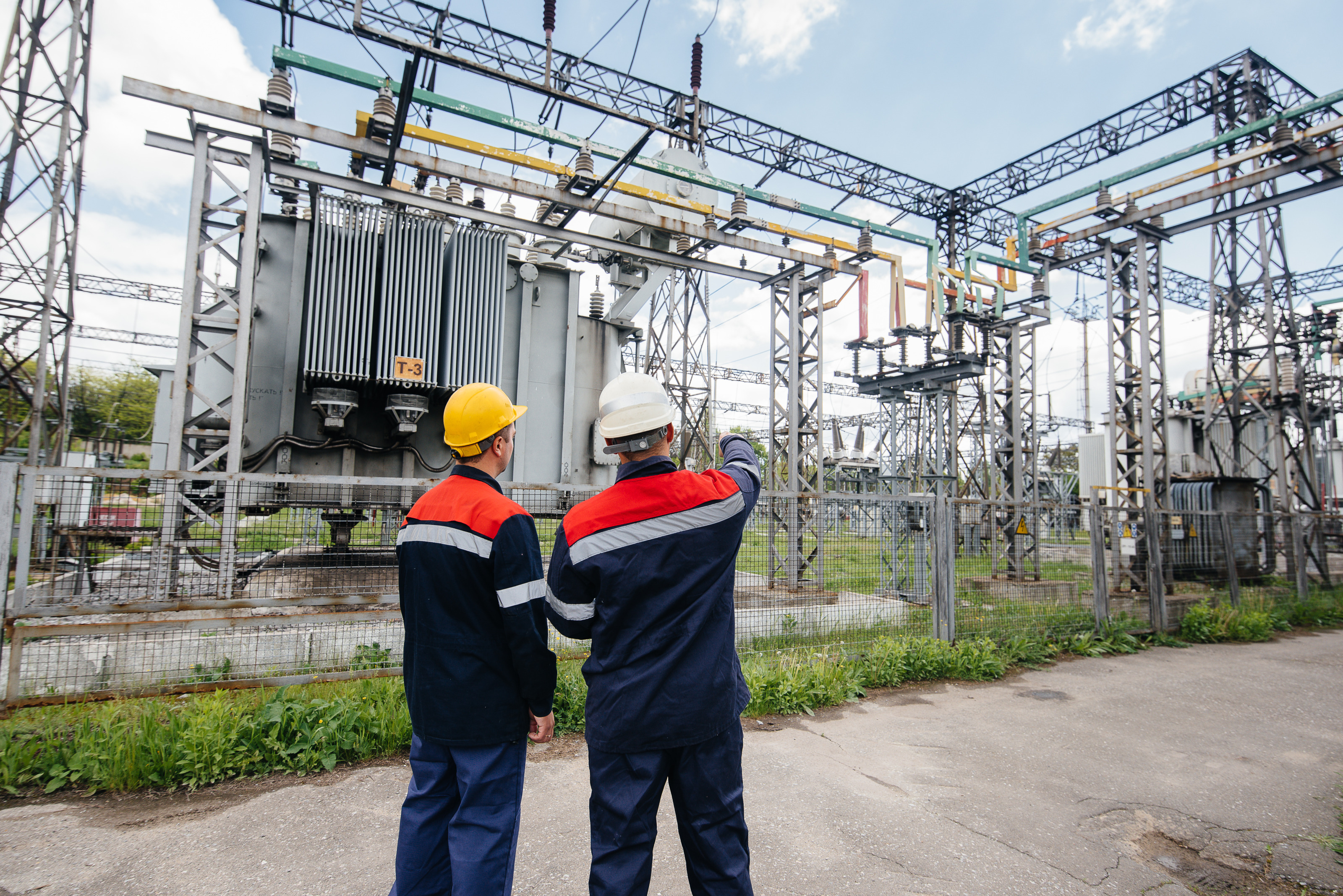 Zwei Personen in Arbeitskleidung und Schutzhelmen stehen vor einem umz&auml;unten Umspannwerk. Eine Person hebt den Arm und zeigt auf Bauteile der Anlage. Im Hintergrund sind Transformatoren, Leitungen, Isolatoren und Metallkonstruktionen zu sehen