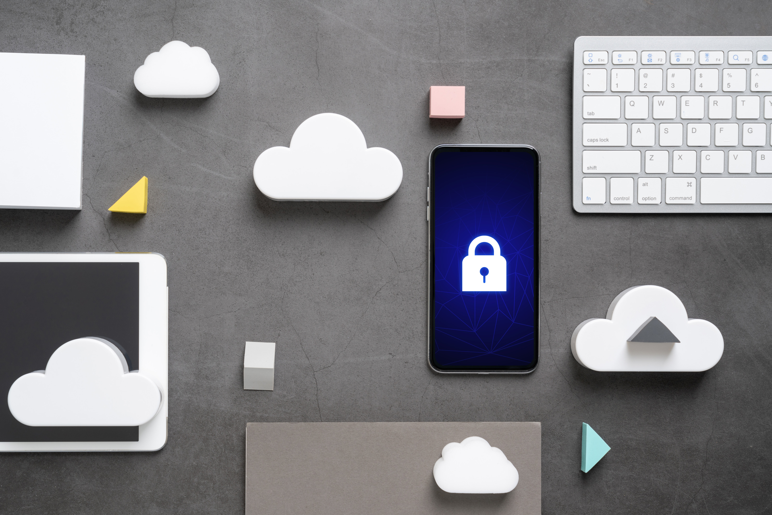 Mehrere Gegenst&auml;nde nebeneinander auf grauem Hintergrund drapiert: Wei&szlig;e Computertastatur, Smartphone auf dessen Display wei&szlig;es Sicherheitsschloss leuchtet, wei&szlig;e Modellwolken aus Schaumstoff, Tablet und weitere Formen aus Schaumstoff