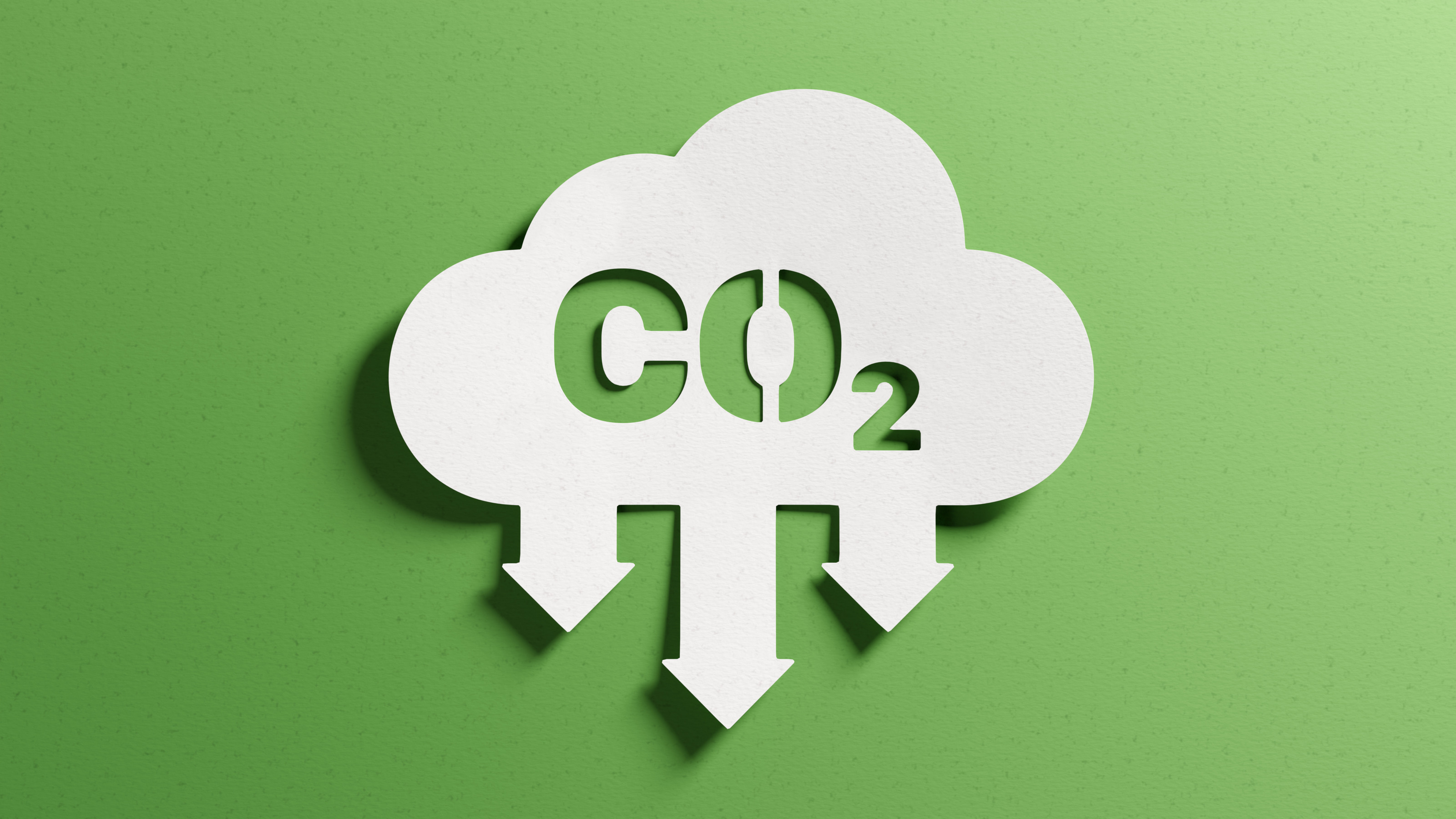 Weiße Wolke aus Papier mit nach unten kragenden Pfeilen, im Inneren Schriftzug CO2 ausgeschnitten, auf grünem Hintergrund