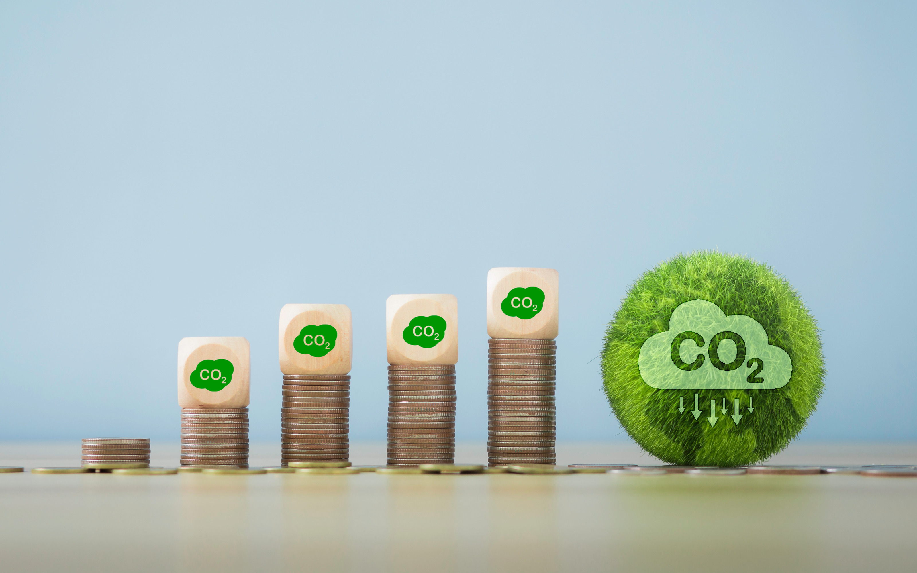 Fünf in der Höhe steigende Münztürmchen mit vier Holzwürfeln obenauf mit der Aufschrift CO2 nebeneinander platziert, daneben Kugel mit Grasoberfläche und darin befindlicher Wolke mit Aufschrift CO2 und Pfeilen nach unten