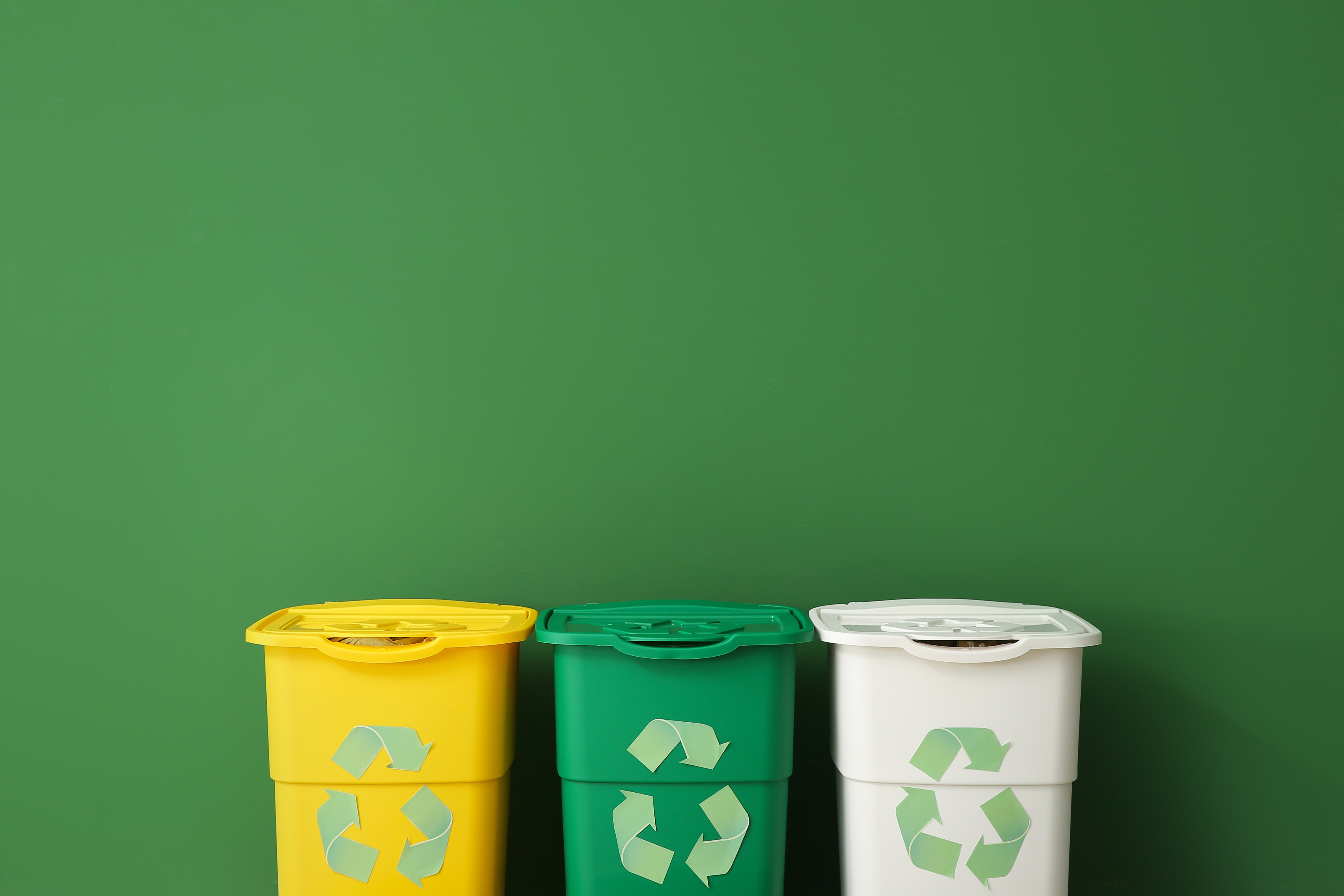 Vor einer gr&uuml;nen Wand stehen nebeneinander drei M&uuml;lltonnen, eine gelbe, eine gr&uuml;ne und eine wei&szlig;e. Auf jeder Tonne ist ein gr&uuml;nes Dreieck, das aus drei Pfeilen besteht