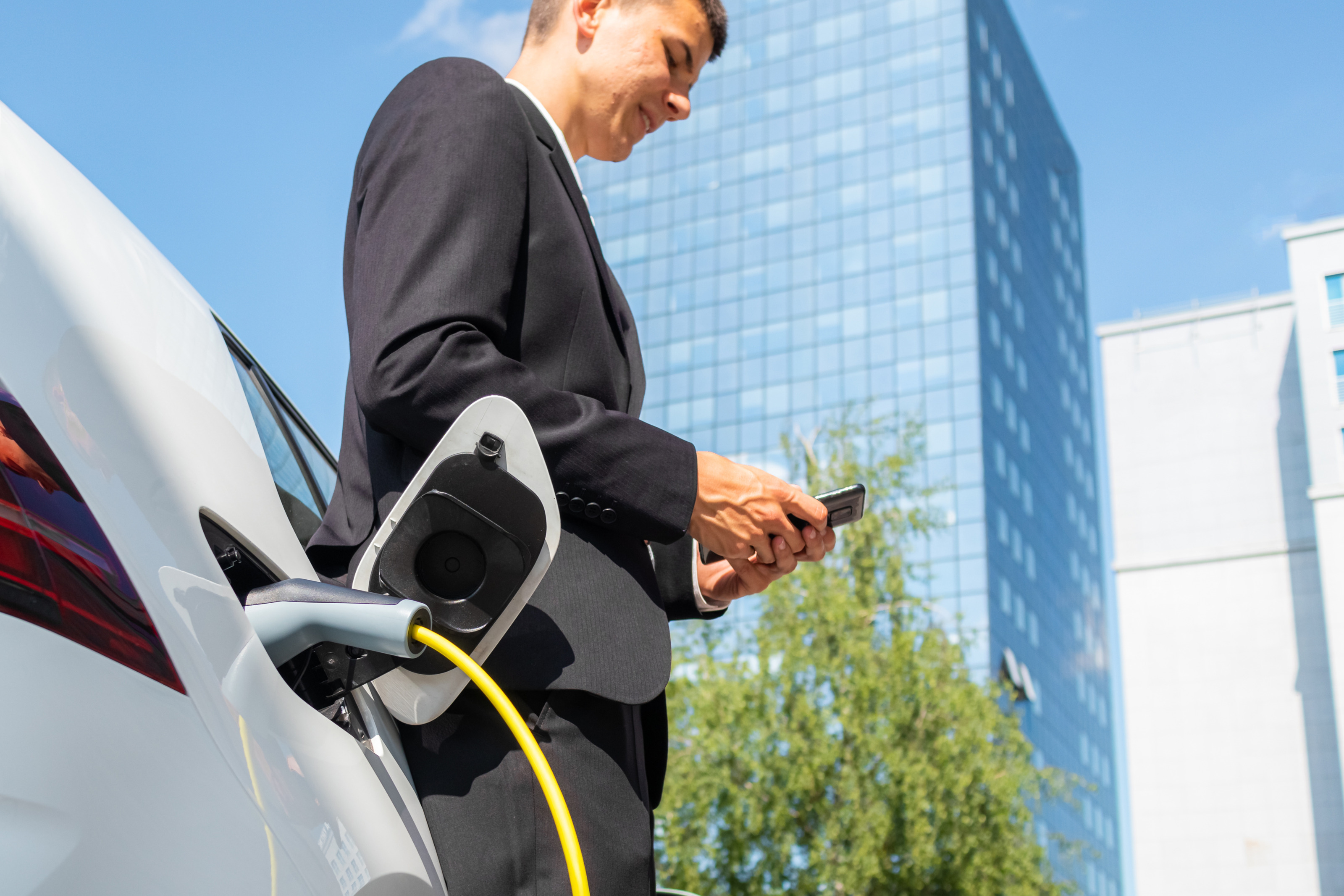 Eine Person in einem Anzug lehnt sich an einem geparkten, wei&szlig;en Auto an und blickt auf ein Handy, das sie h&auml;lt. Das Auto ist an einer E-Ladestation angeschlossen. Im Hintergrund ist ein Hochhaus