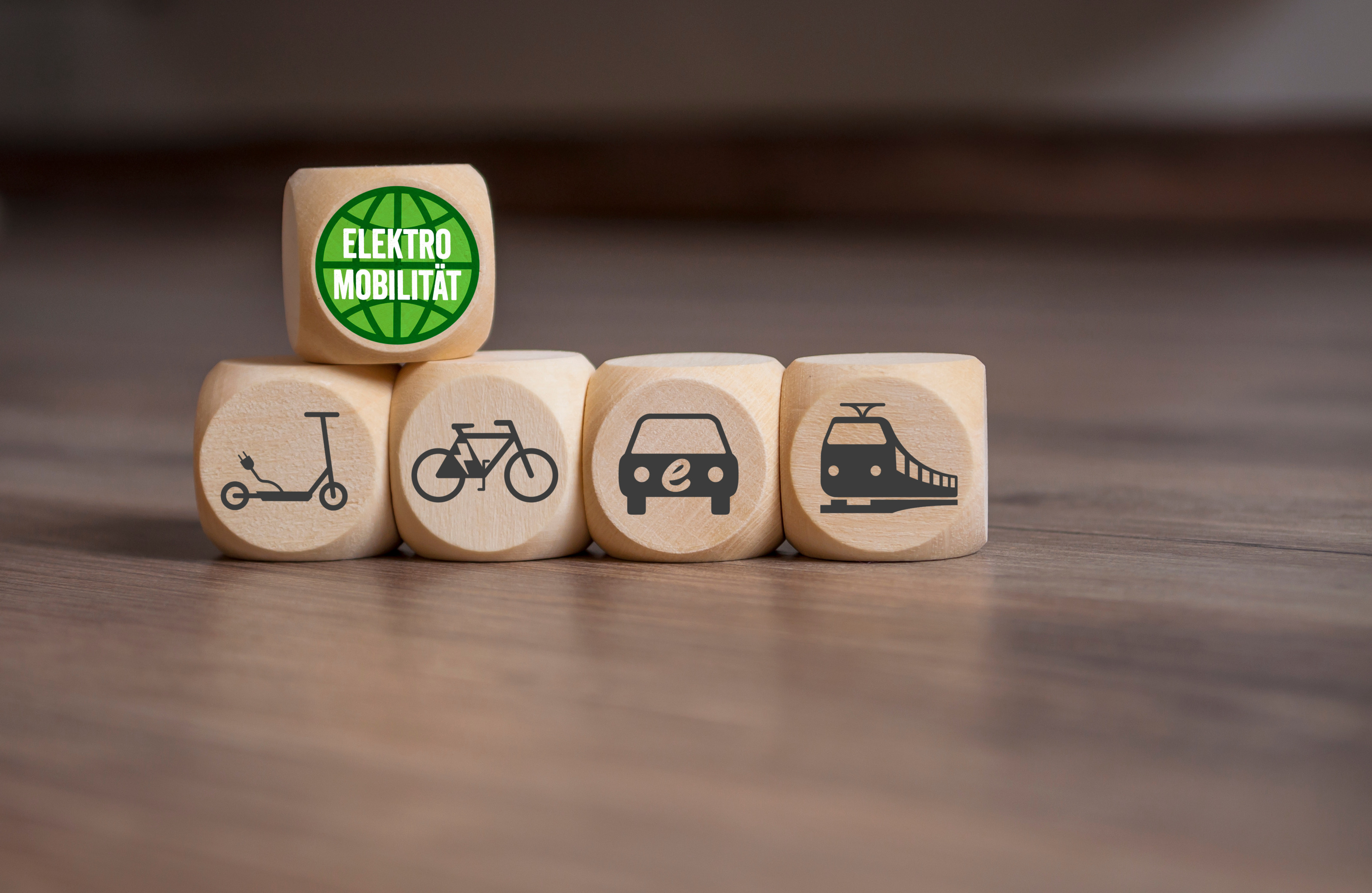 Vier Holwürfel mit Symbolen E-Roller, Fahrrad, Elektroauto und Zug nebeneinander platziert, oben auf ein weiterer Holzwürfel mit weißem Schriftzug Elektromobilität in grünem Kreis
