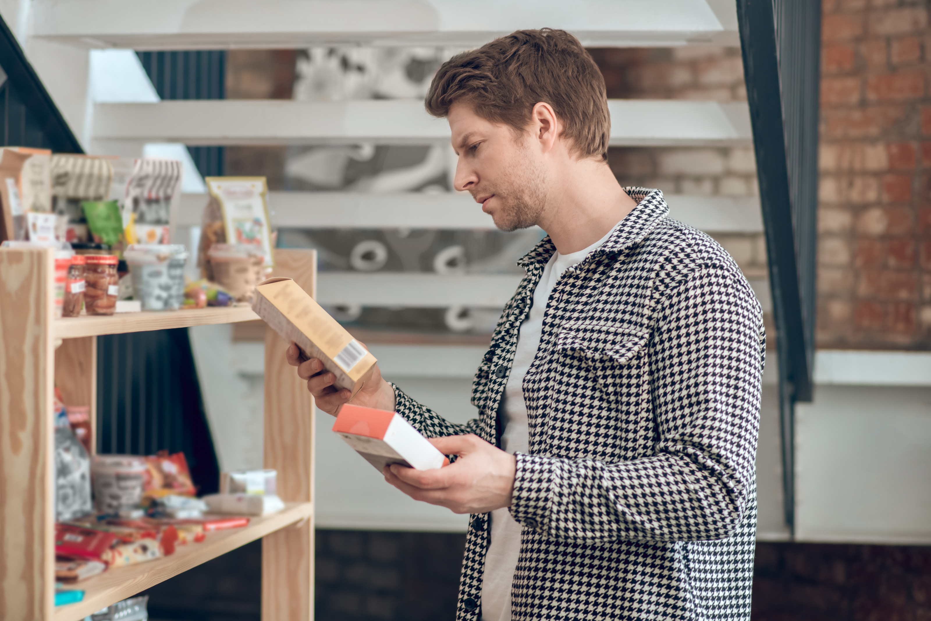 Eine Person steht neben einem Holzregal. Im Regal stehen verschiedene Lebensmittel. Die Person h&auml;lt in beiden H&auml;nden jeweils eine Verpackung