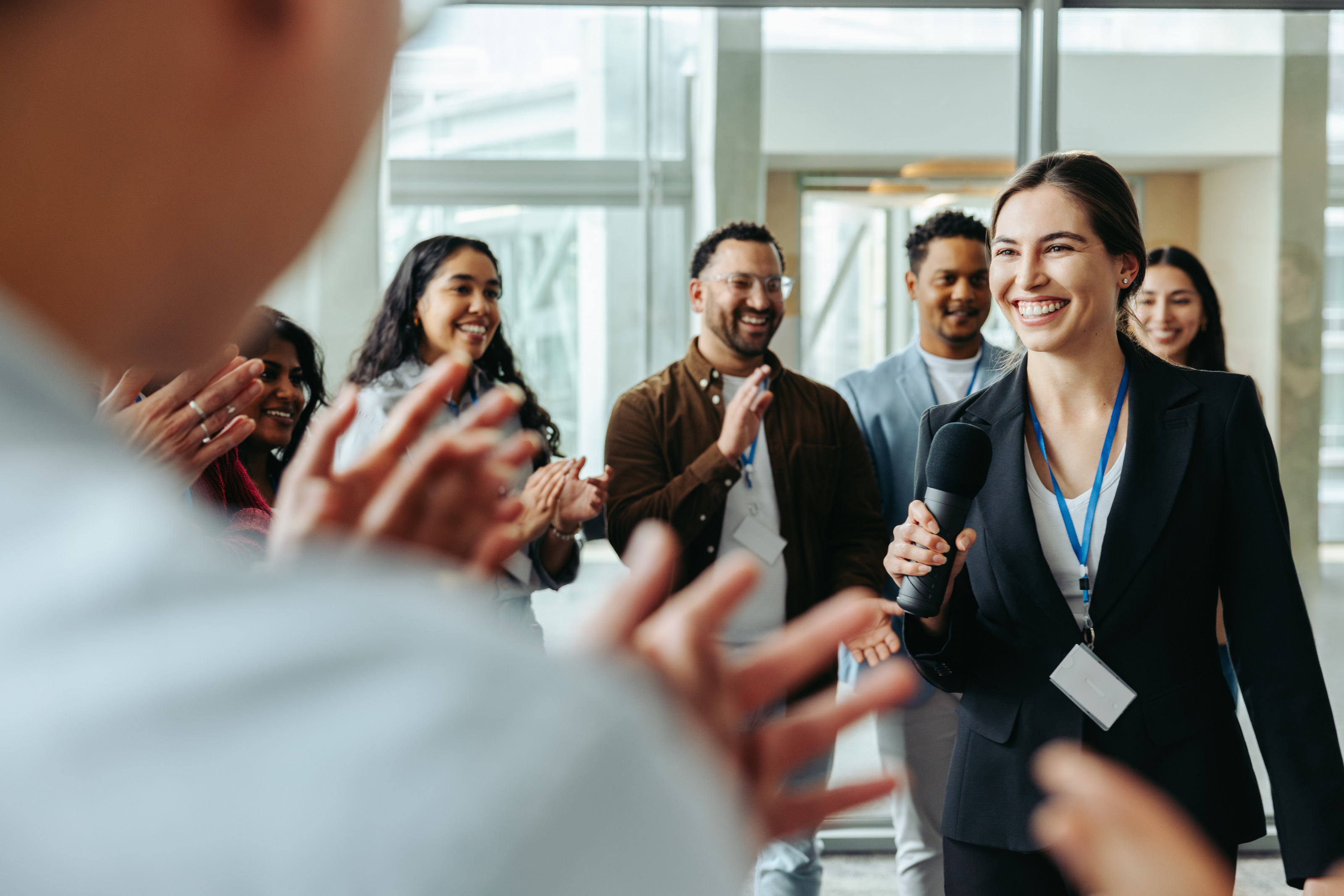 Personen mit Ausweisschildern stehen in einem Halbkreis und klatschen. Im Kreis steht eine Person mit dunklem Sakko, Mikrofon sowie Ausweisschild, die l&auml;chelt