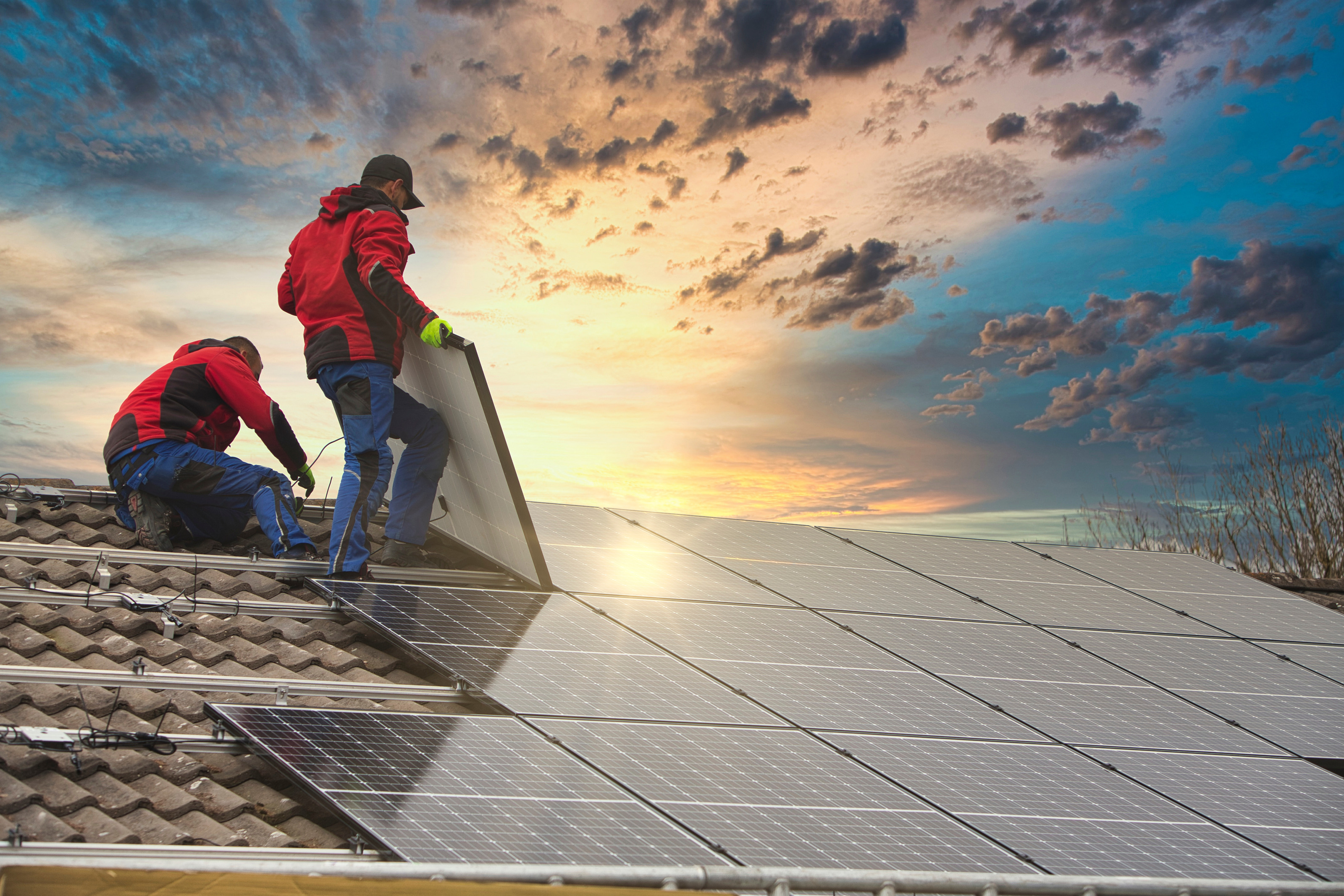 Zwei Personen mit Handschuhen und Arbeitskleidung stehen nebeneinander auf einem schr&auml;gen Dach und installieren auf diesem Solarkollektoren. Hinter ihnen ist der bew&ouml;lkte Himmel