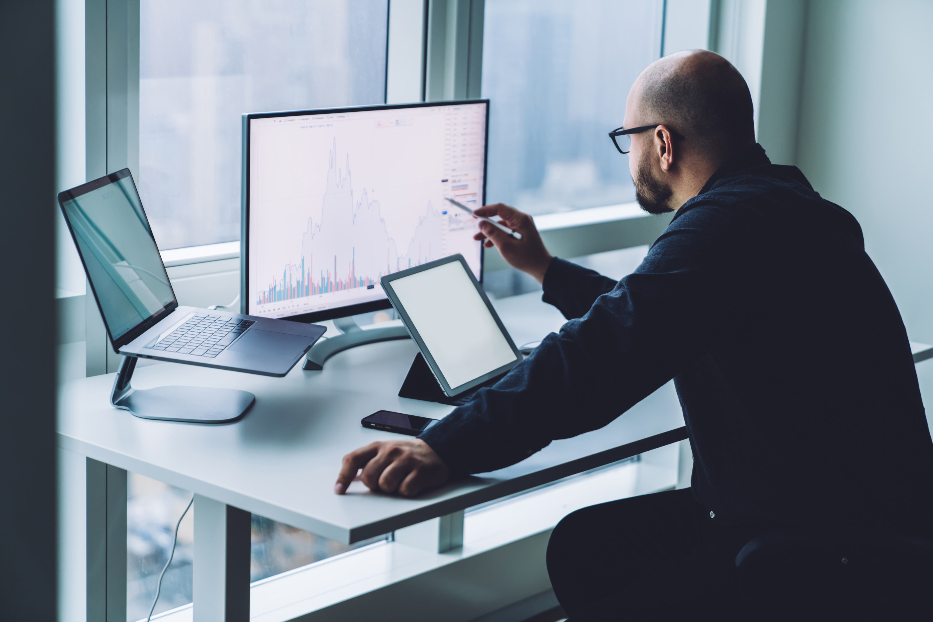 Eine Person in R&uuml;ckenansicht sitzt vor einem Tablet, einem Laptop und Computerbildschirm mit Diagrammen und arbeitet.