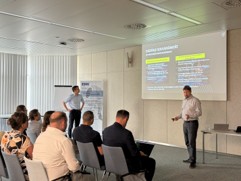 Das Foto zeigt einen Seminarraum mit mehreren sitzenden Personen, die einer Pr&auml;sentation folgen. Vor einer Projektionsfl&auml;che steht eine Person und pr&auml;sentiert Folien
