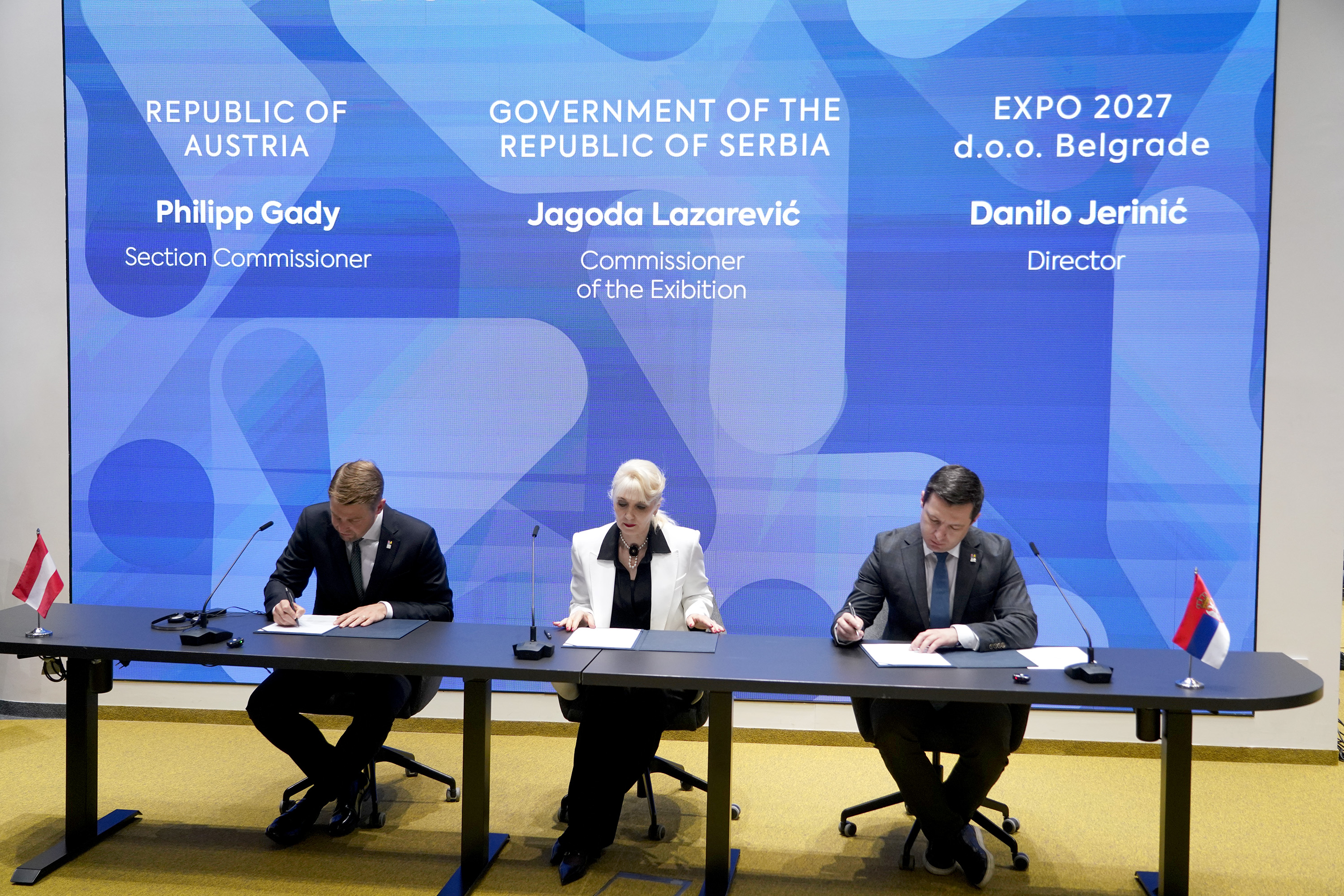 Drei Personen unterzeichnen Dokumente vor Hintergrund mit Text zu &Ouml;sterreich, Serbien und Expo 2027