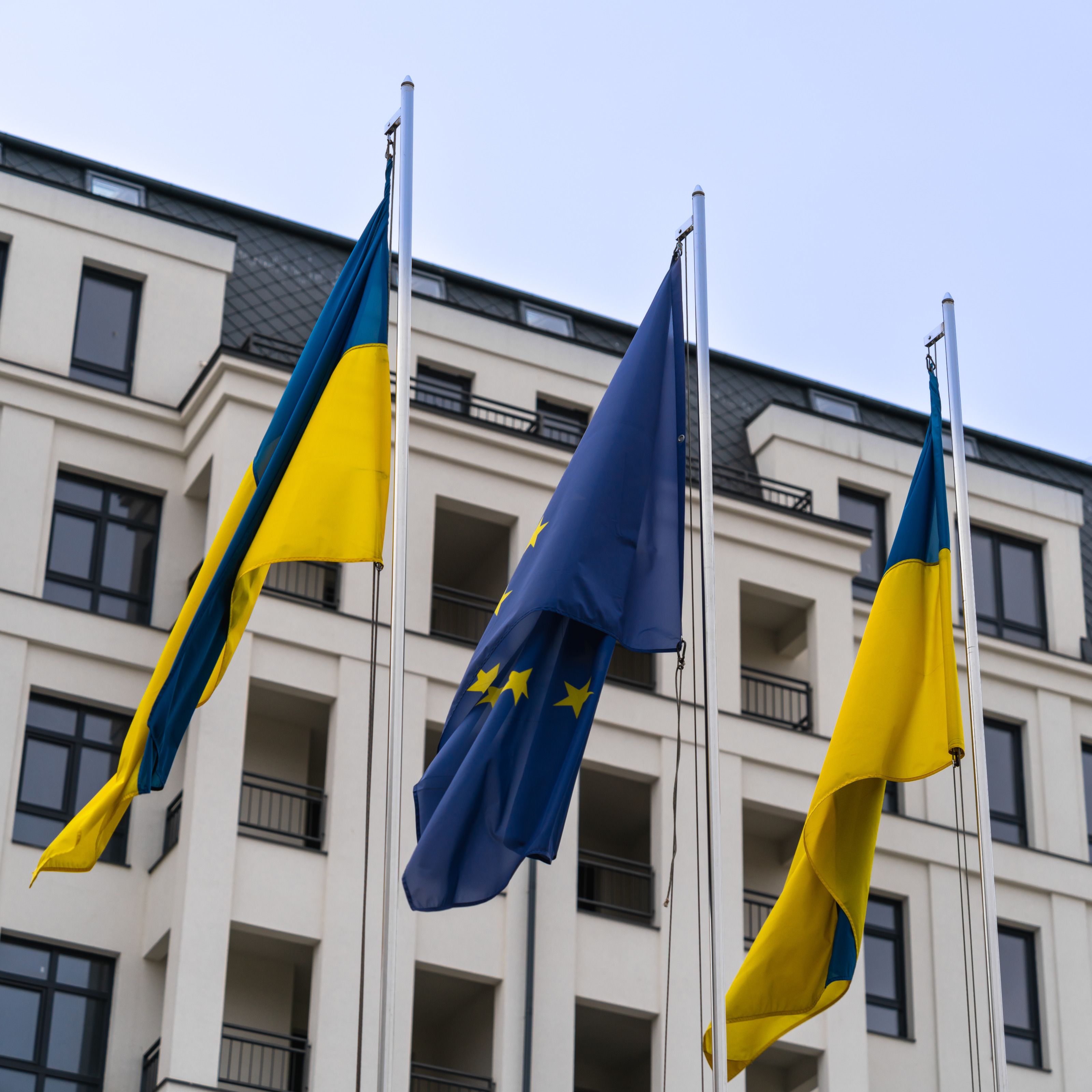 Drei Flaggen an Masten in Wind vor modernem Geb&auml;ude im Hintergrund: die beiden &auml;u&szlig;eren Flaggen der Ukraine, blau-gelb gestreift, in der Mitte dazwischen die EU-Flagge - blauer Stoff mit gelben in Kreisform verlaufenden Sternen