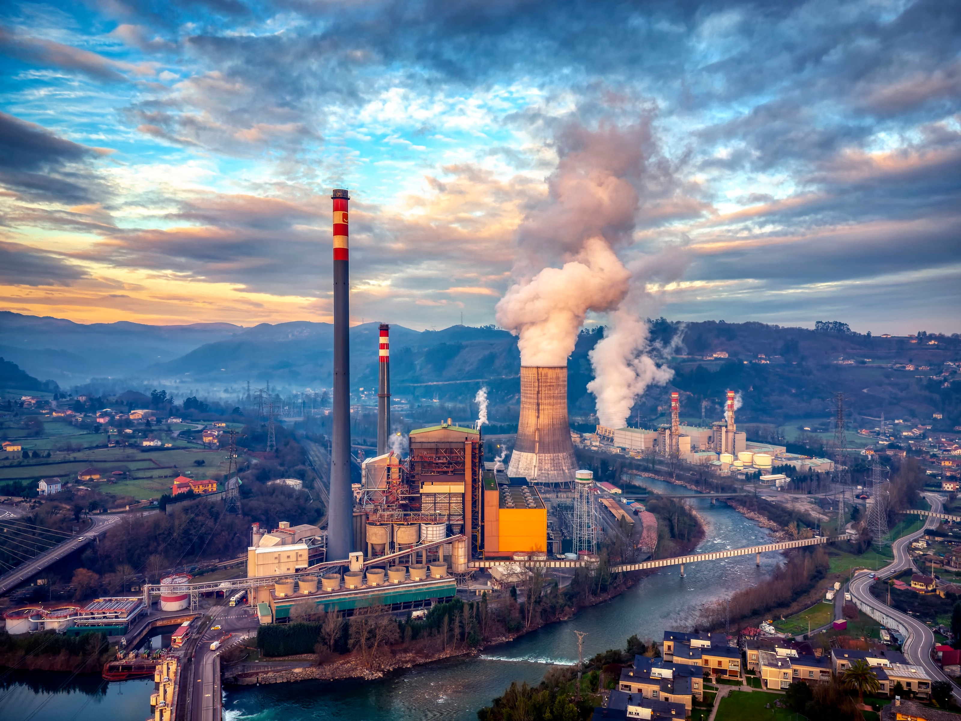 An einem Fluss liegen links und rechts Fabrikgel&auml;nde. Aus einigen Schornsteinen und T&uuml;rmen steigt Rauch auf. Neben der Fabrik sind weitere Geb&auml;ude, Stra&szlig;en, Wiesen und H&uuml;gel
