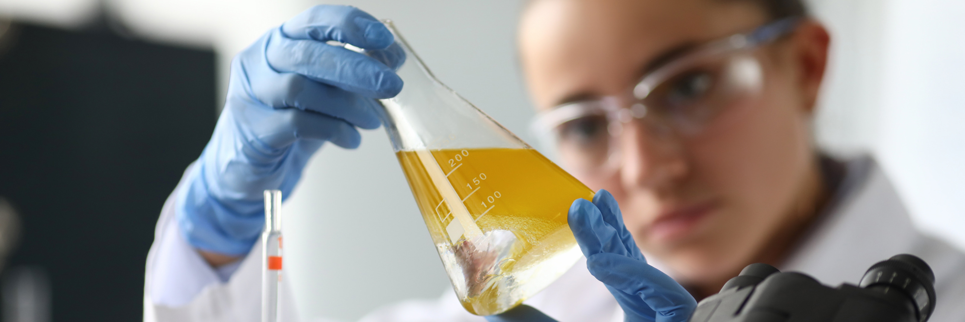 Im Fokus Laborglas mit gelber Fl&uuml;ssigkeit, das Person mit Schutzbrillen verschwommen im Hintergrund neigt und darauf blickt