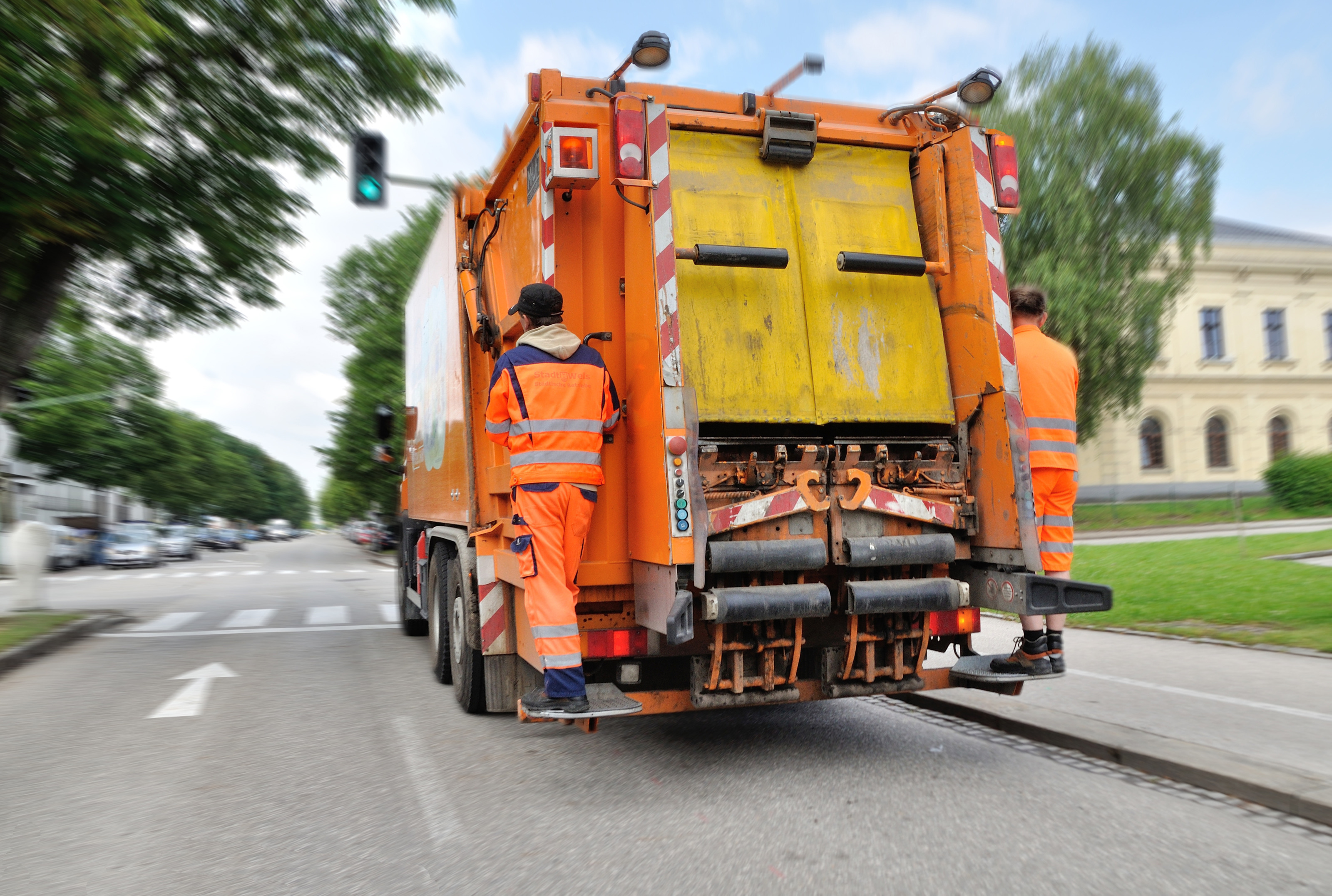 Zwei Personen in orange-reflektierenden Jacken und Hosen stehen hinten auf einem M&uuml;llauto und halten sich fest. Das M&uuml;llauto f&auml;hrt auf einer Stra&szlig;e. Im Hintergrund sind parkende Autos, Geb&auml;ude und eine Allee.