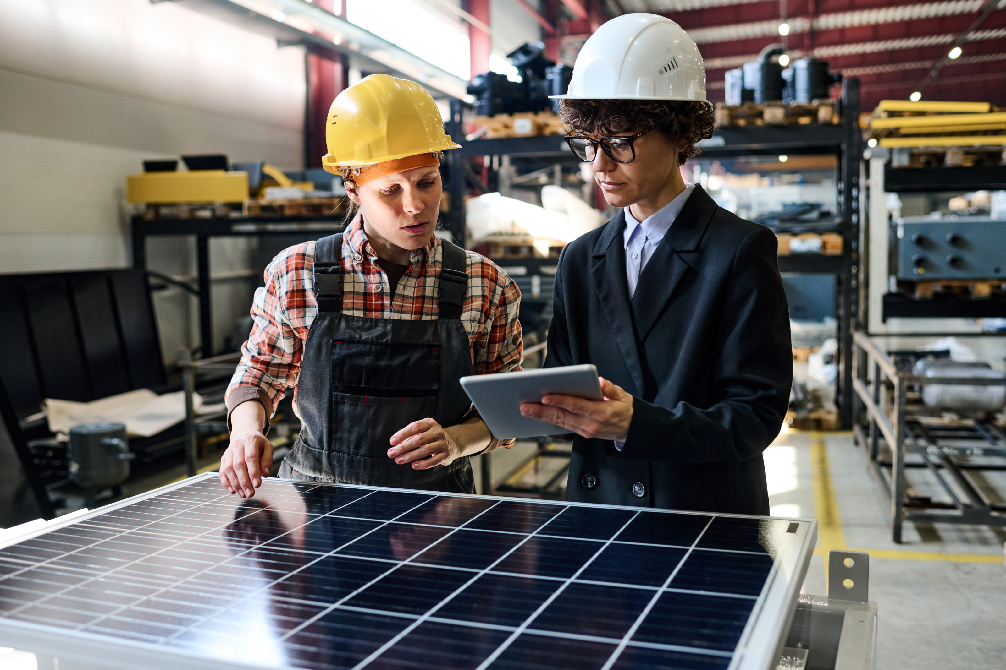 Zwei Personen mit Schutzhelmen blicken auf Tablet, das eine Person in H&auml;nden h&auml;lt, vor ihnen Solarpanel, im Hintergrund verschwommen Regale und Maschinen