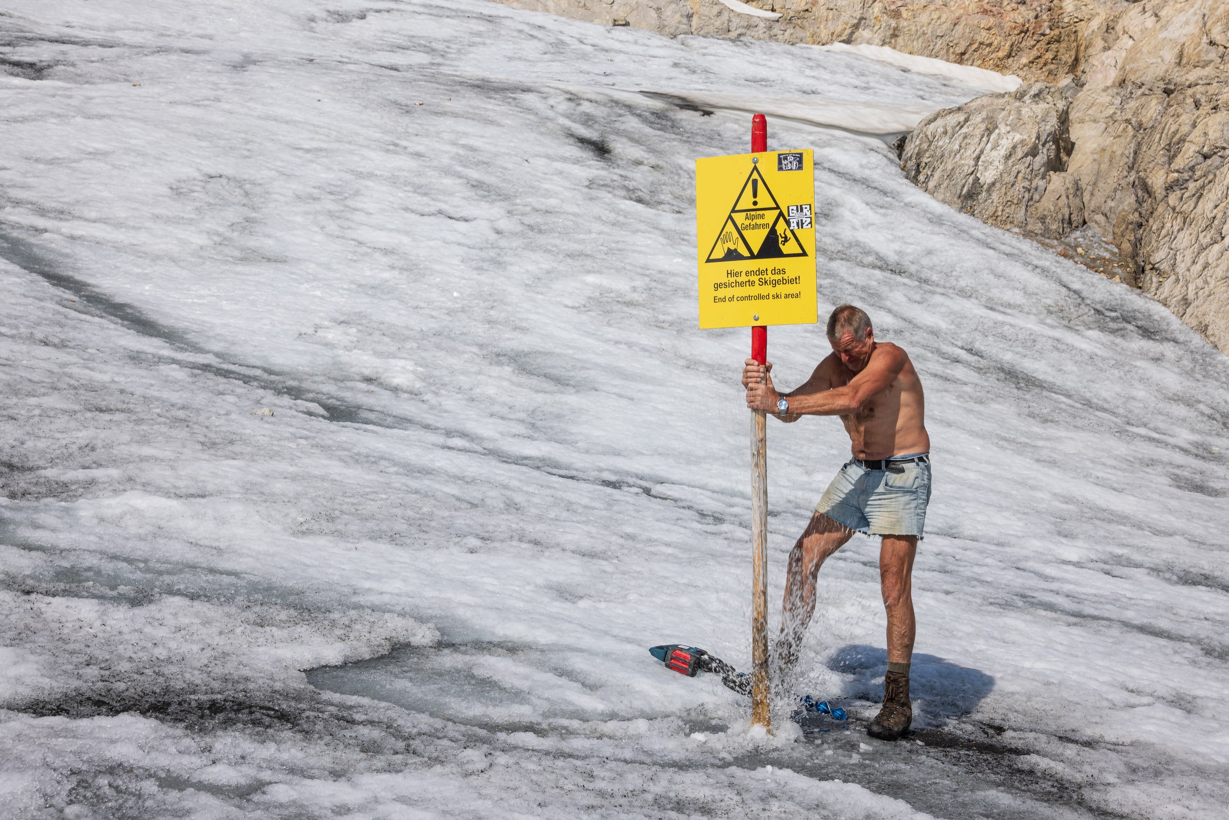 Das Bild zeigt einen leicht bekleideten Arbeiter, der ein auf einem Holzpfahl befestigtes Schild mit der Aufschrift &bdquo;Hier endet das gesicherte Skigebiet&ldquo; ins schmelzende Eis rammt, w&auml;hrend Schmelzwasser aus dem Boden spritzt.