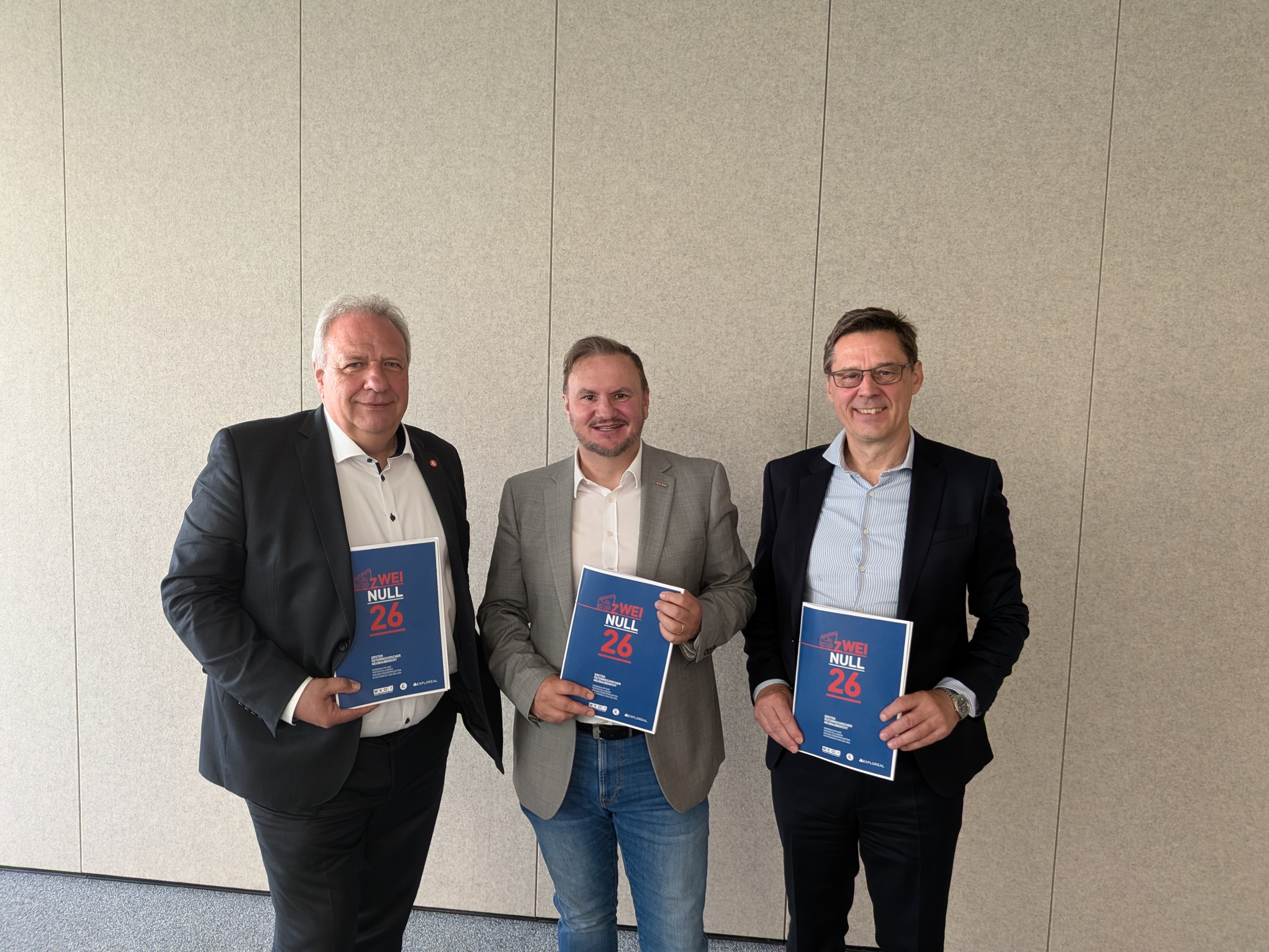 Drei Personen stehen nebeneinander vor einer hellen, neutralen Wand. Jede Person h&auml;lt eine blaue Brosch&uuml;re mit roter und wei&szlig;er Schrift