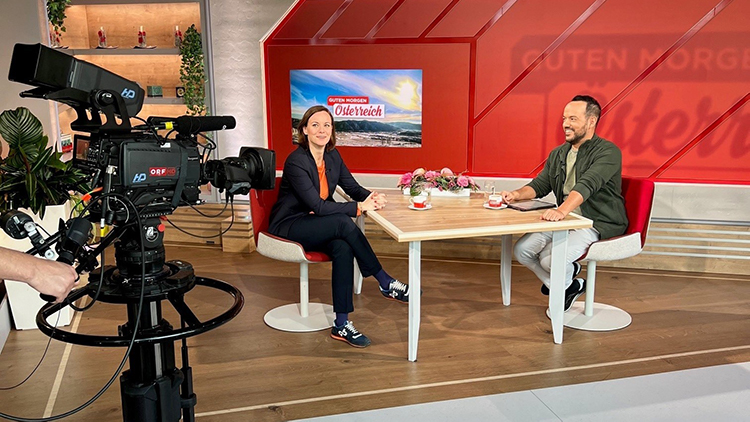 Kamerastill zweier Personen an einem Tisch in Filmstudio sitzend, im Vordergrund ausschnitt einer Kamera