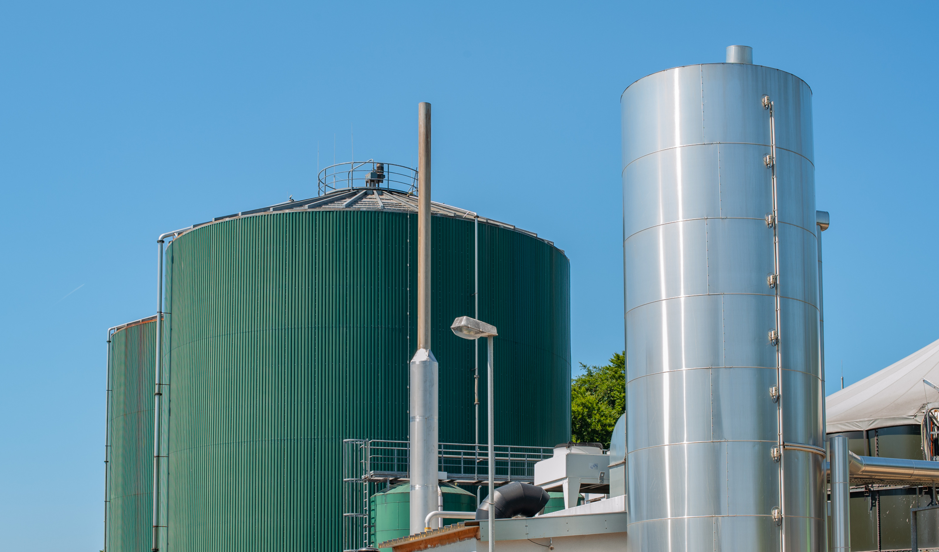 Außenansicht einer Biogasanlage: Große grüne Metallsilos neben silbernen Metallturm unter blauem Himmel