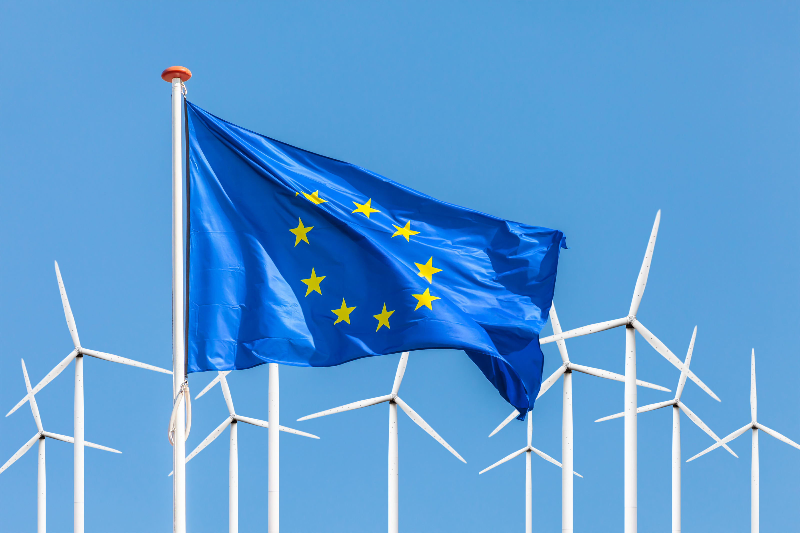 EU-Flagge im Wind wehend, im Hintergrund Windräder unter blauem Himmel