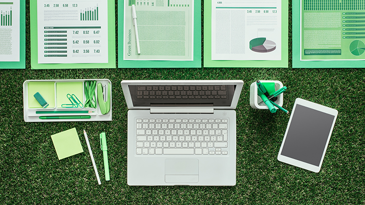 Vogelperspektive auf aufgeklapptem Laptop auf Rasen platziert umgeben von Dokumenten und Büroutensilien in grün