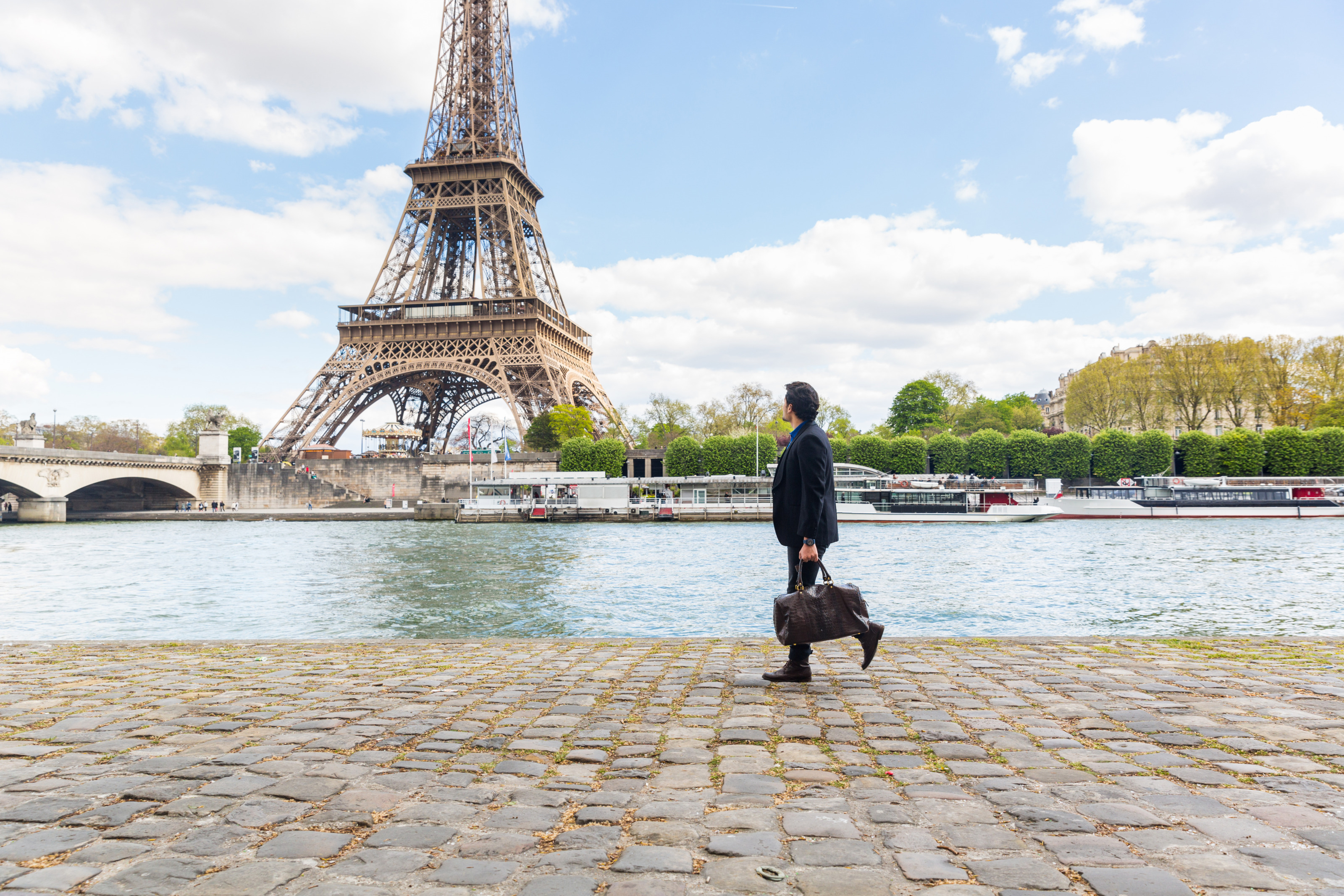 Eine Person in Sakko und mit Ledertasche geht an einer Uferpromenade entlang. Auf dem Fluss neben ihr sind lange Boote. Im Hintergrund sind eine Br&uuml;cke und der Eiffelturm