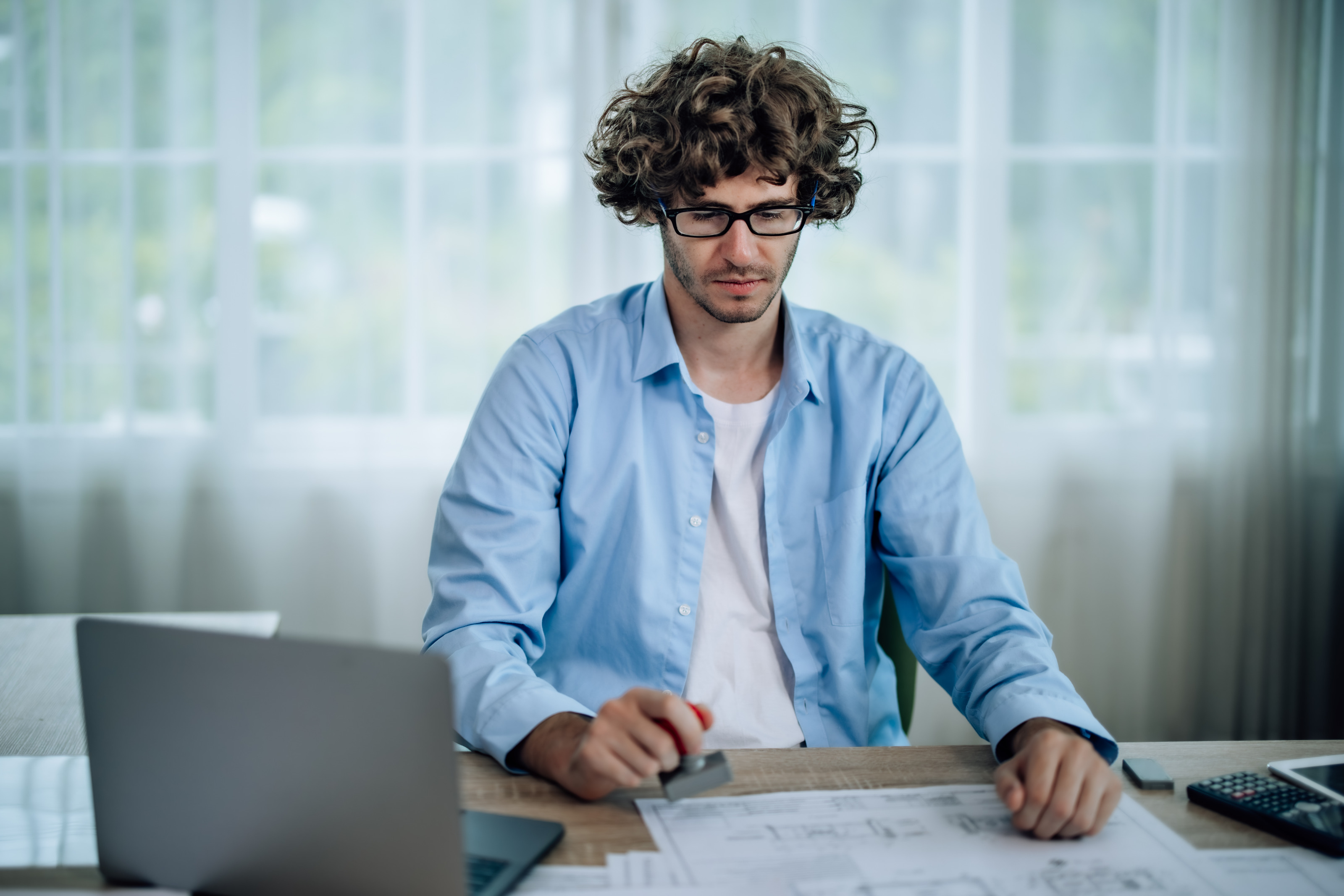 Person mit Brillen in blauem Hemd sitzt an Schreibtisch vor aufgeklapptem Laptop und stempelt vor sich liegenden Plan