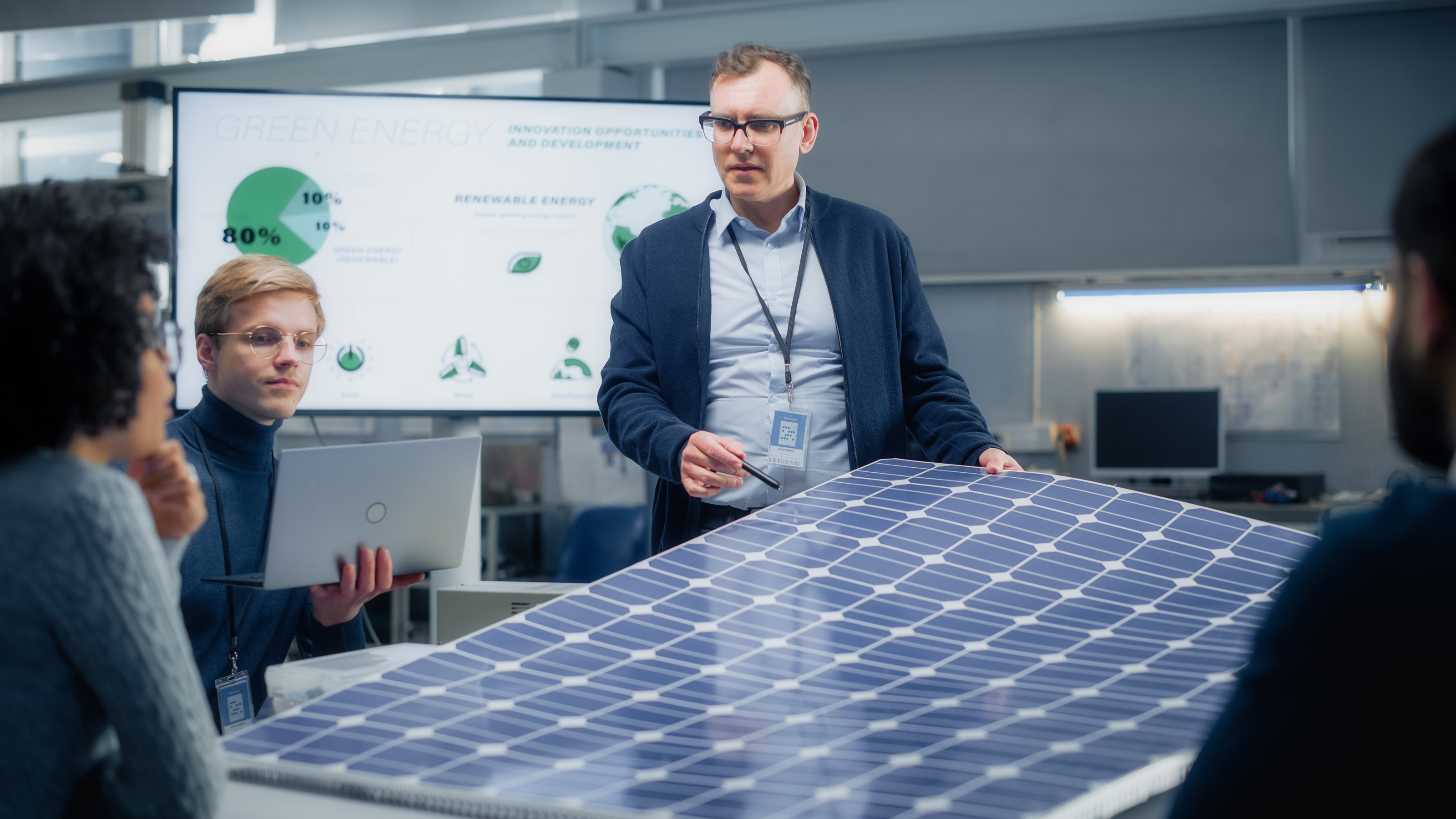 Vier Personen sitzen bzw. stehen um einen Tisch. In der Mitte liegt eine große Photovoltaikoberfläche. Hinter den Personen ist ein großer Bildschirm, auf dem green energy steht.