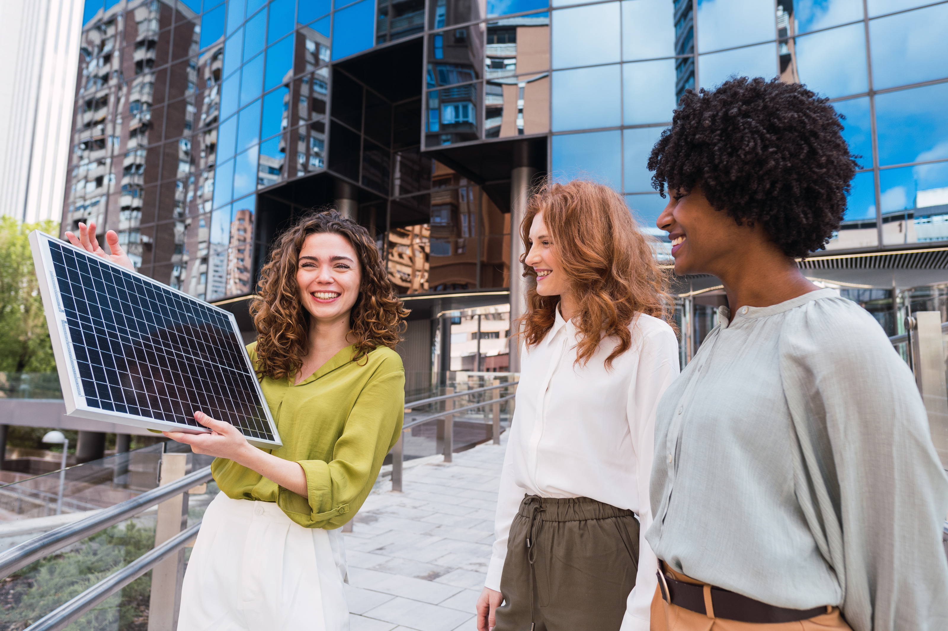 Drei lächelnde Personen stehen vor modernem Gebäude, eine Person hält Solarpanel in Händen