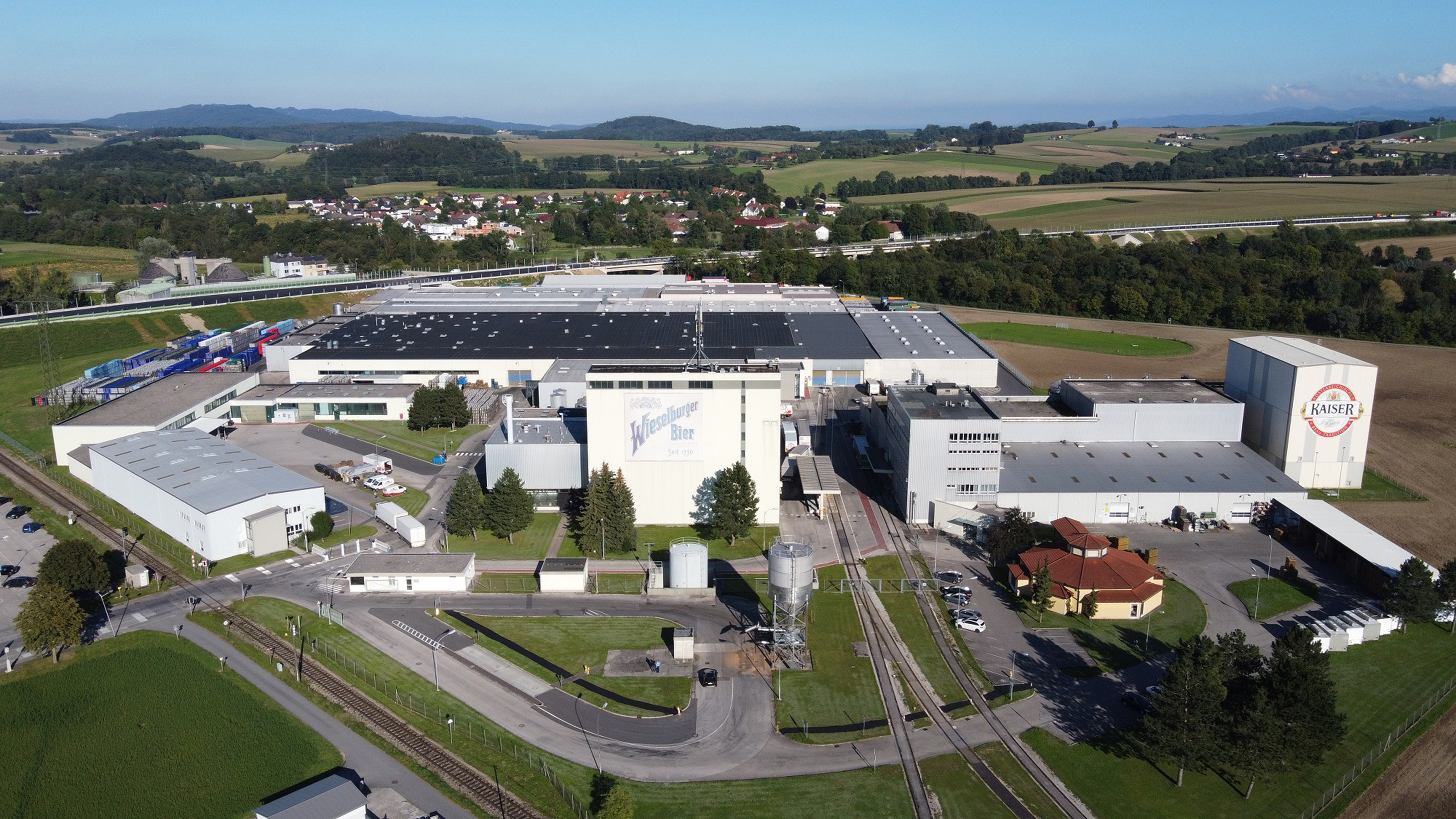 Luftansicht der Brauerei Wieselburg: Großes Industrieareal mit Hallen bebaut umgeben von grünen Wiesen, Bäumen und Autobahn