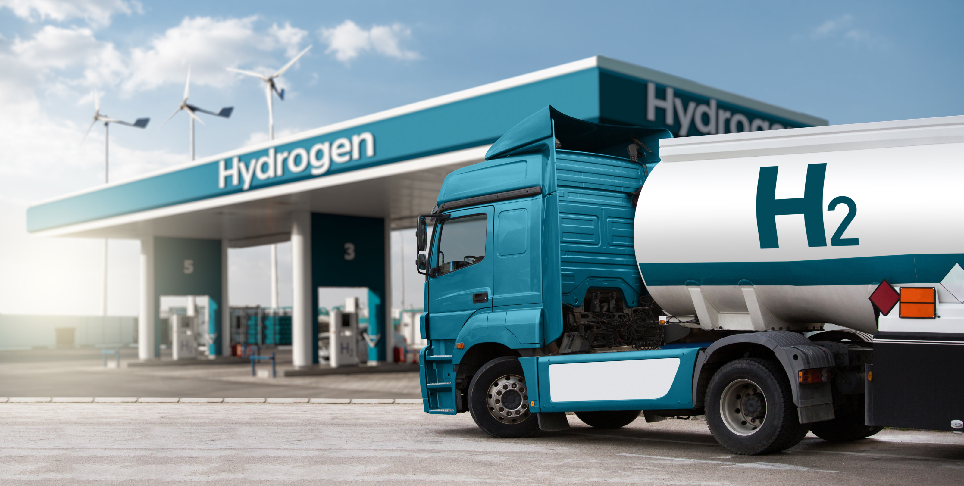 Blauer Lastkraftwagen mit weißem Tankanhänger mit der Aufschrift H2 für Wasserstoff steht vor Tankstelle mit Aufschrift Hydrogen, im Hintergrund drei Windräder