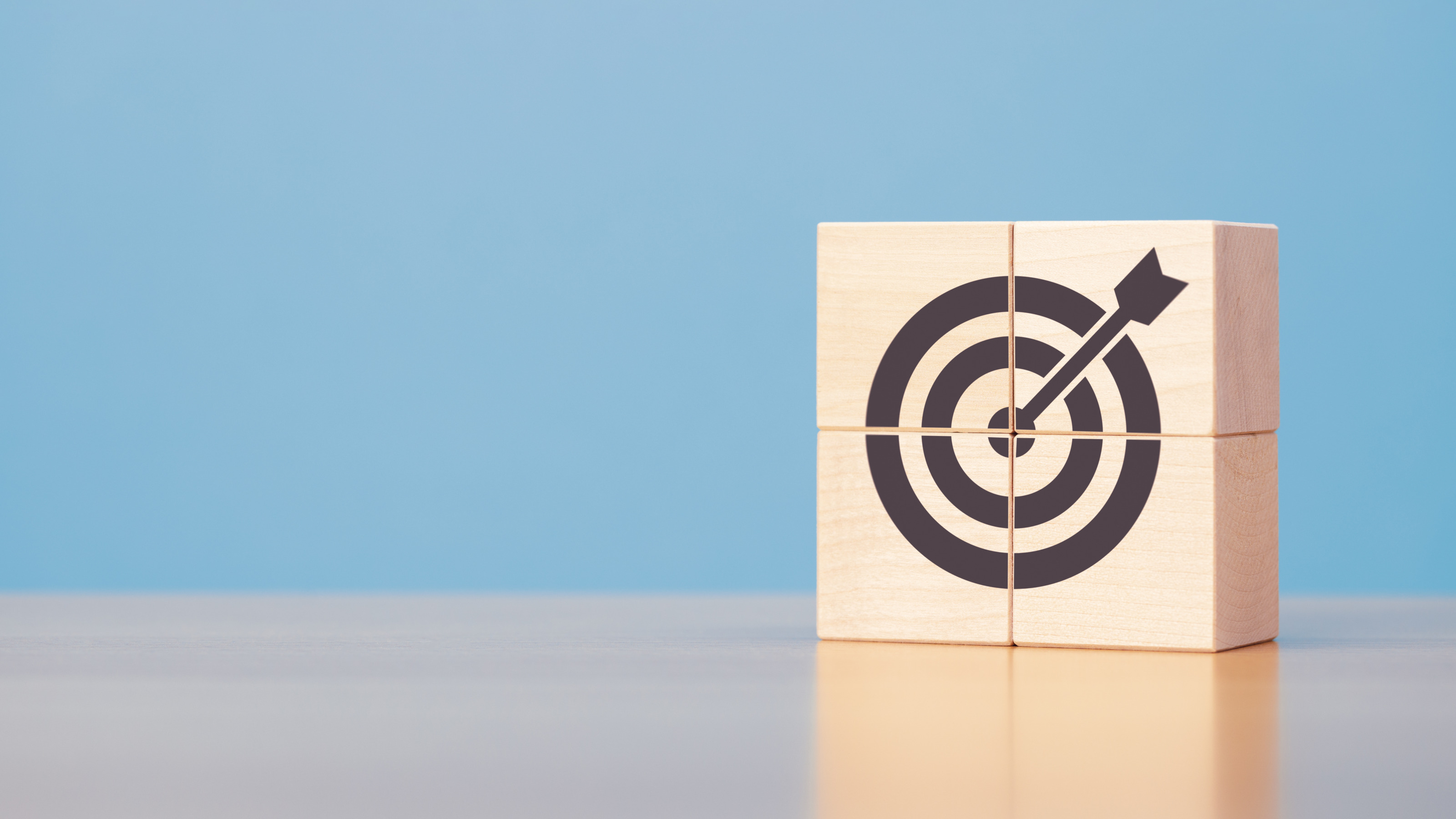 Vor blauem Hintergrund auf einem Tisch stehen vier W&uuml;rfel, die ein Quadrat ergeben. Auf den W&uuml;rfeln ist eine Zielscheibe