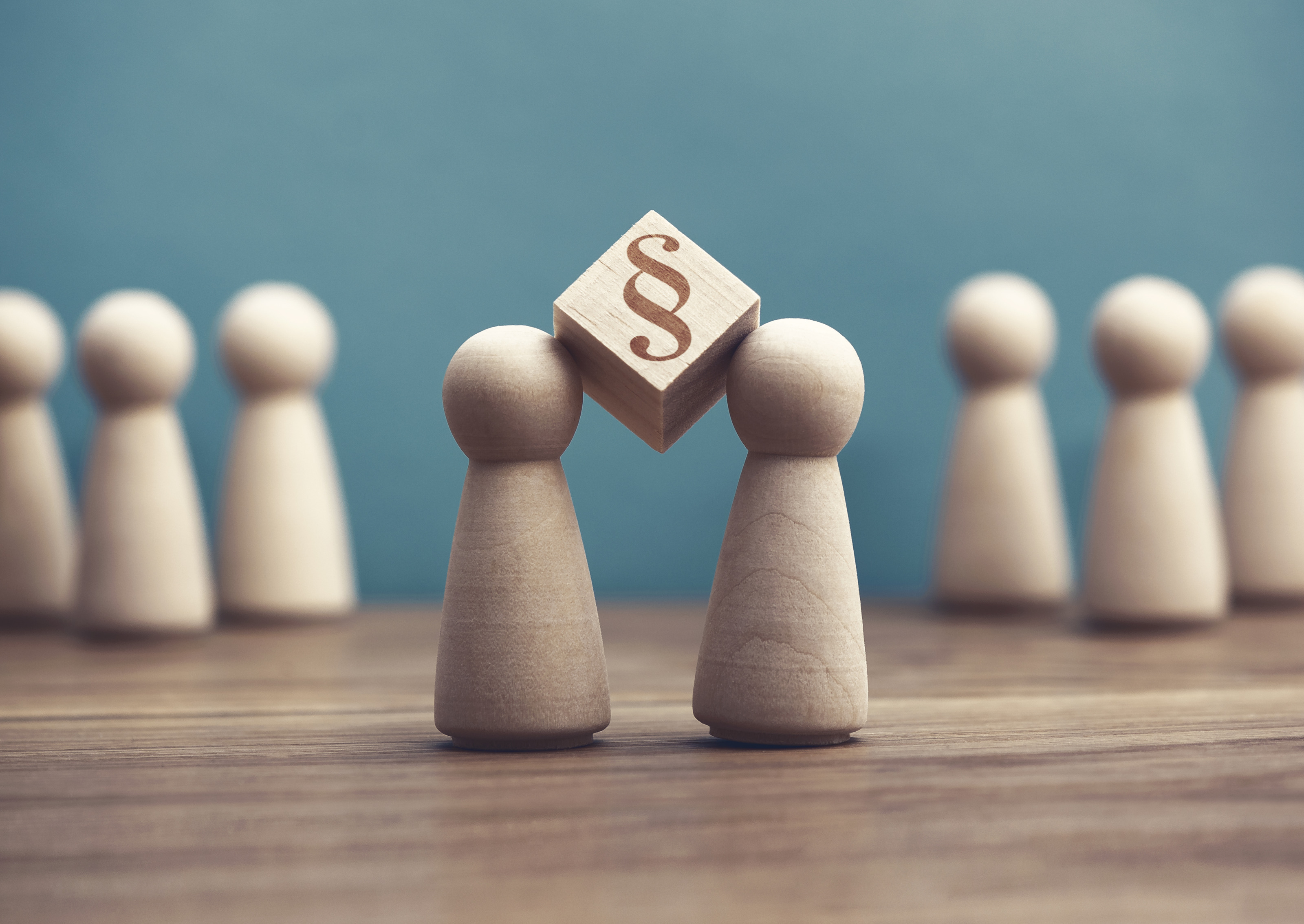 Auf einer hölzernen Oberfläche stehen im Vordergrund zwei kleine Holzfiguren, zwischen deren Köpfen ein Holzwürfel gehalten wird, auf dem ein Paragrafensymbol ist. Im Hintergrund stehen weitere Holzfiguren