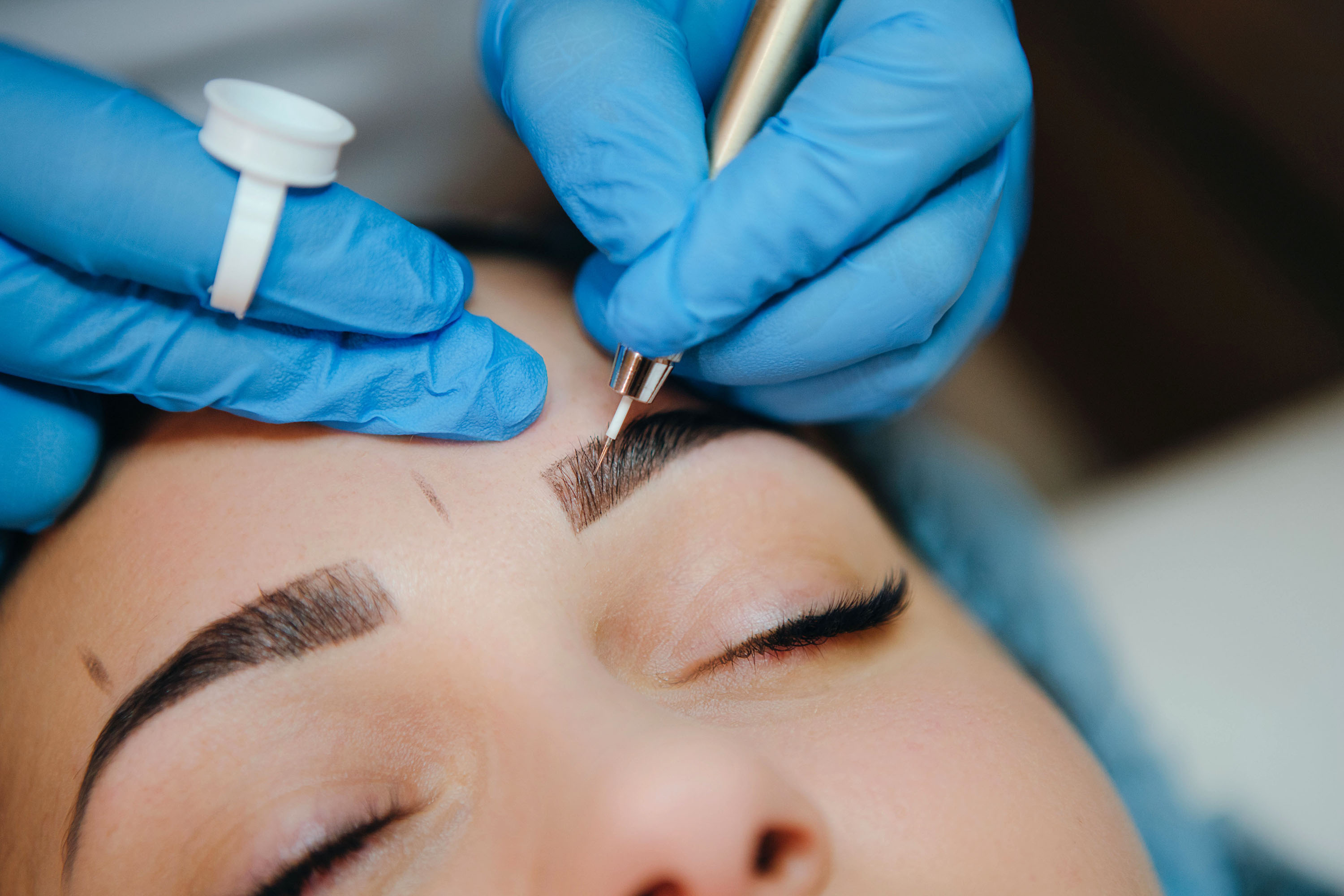 Nahaufnahme einer Microblading-Behandlung.: Eine Person mit geschlossenen Augen bekommt von einer anderen Person mit Gummihandschuhen mithilfe einer kosmetischen Nadel Augenbrauenh&auml;rchen gezeichnet.