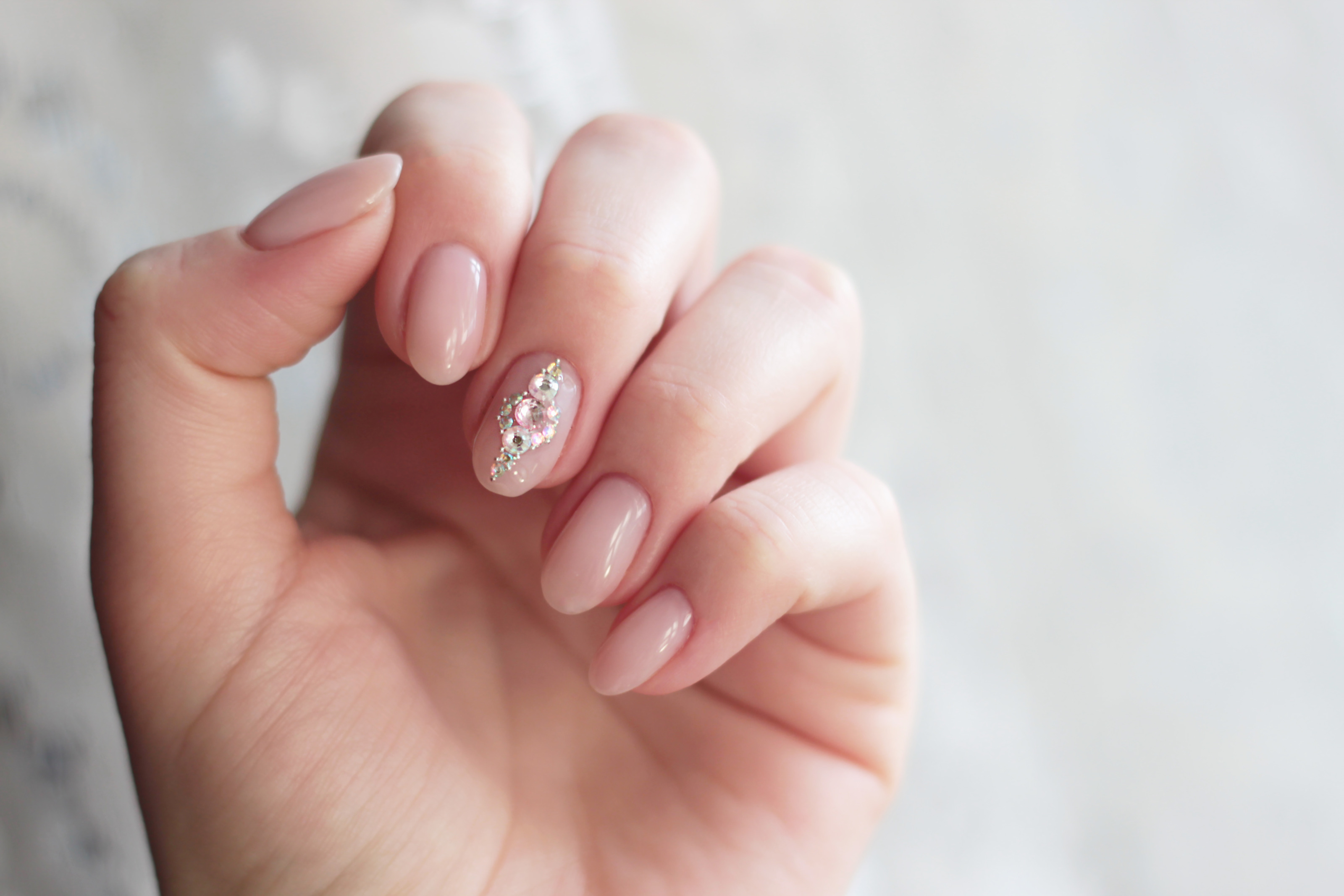 Nahaufnahme von gepflegten, beigen N&auml;geln mit Glitzersteinen am Mittelfinger.