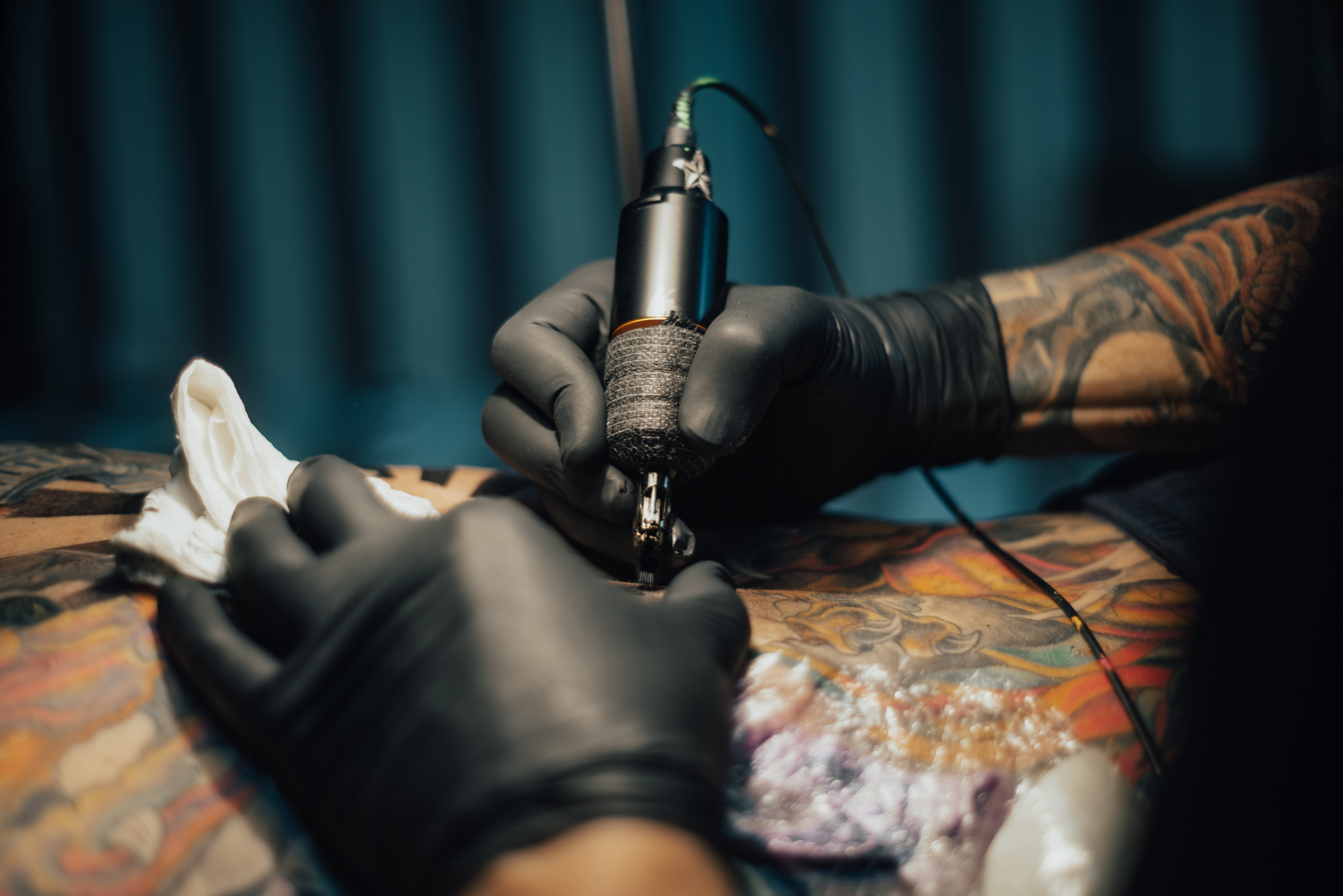 Nahaufnahme eines K&ouml;rperteils einer Person. Die Person liegt. Auf der Haut sind verschiedene Tattoos. Eine andere Person h&auml;lt ihre H&auml;nde auf den K&ouml;rper der liegenden Person. Die rechte Hand h&auml;lt eine T&auml;towiermaschine auf die Haut.