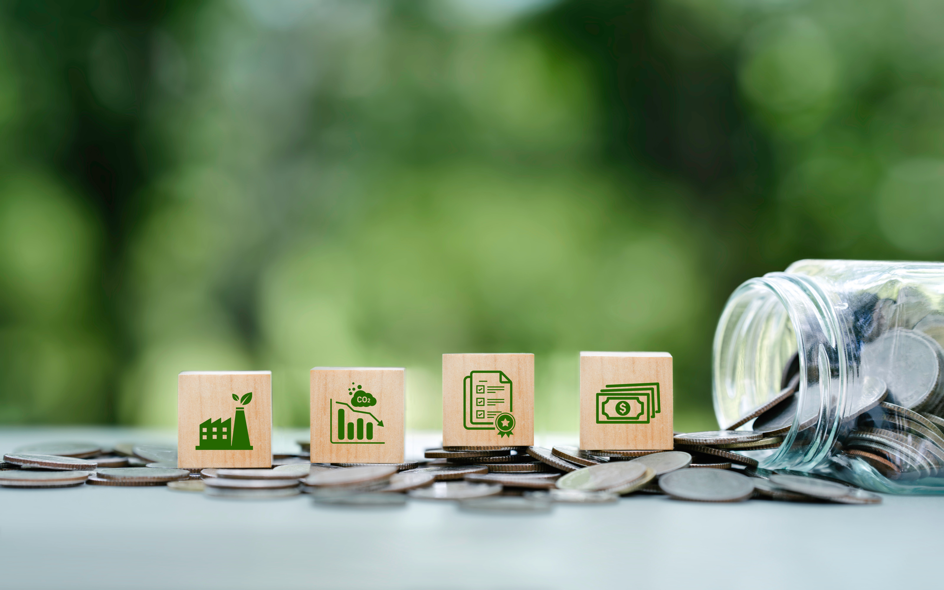 Mehrere Holzwürfel nebeneinander platziert mit Symbolen zum Thema Nachhaltigkeit auf Münzen platziert, nebenliegend umgekipptes Rexglas aus dem Münzen